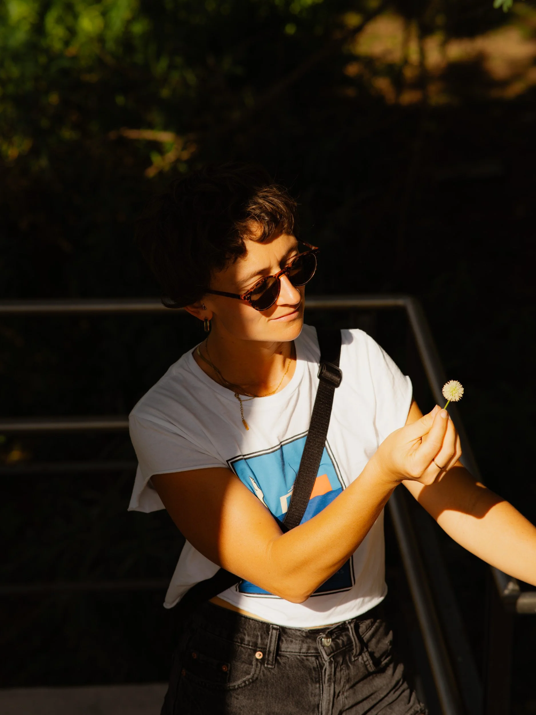 A woman with short dark hair and sunglasses holding a dandelion flower, looking at it thoughtfully while standing outdoors.