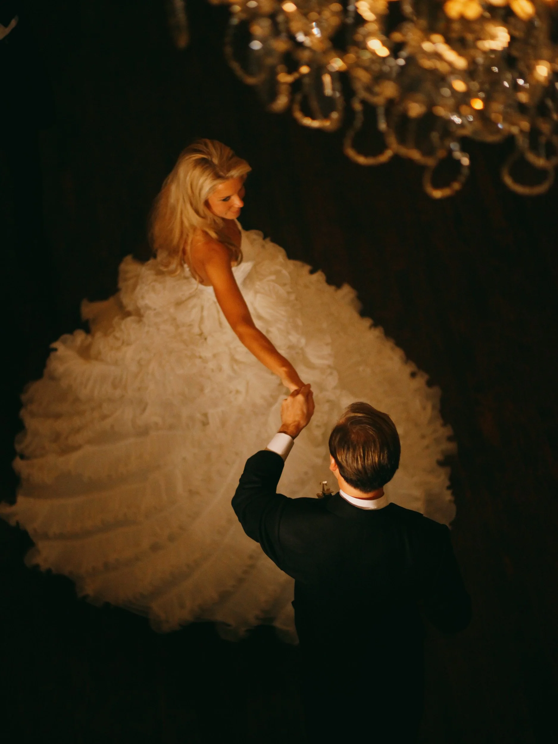 A bride in a white wedding gown dancing with a groom in a black tuxedo under a chandelier.