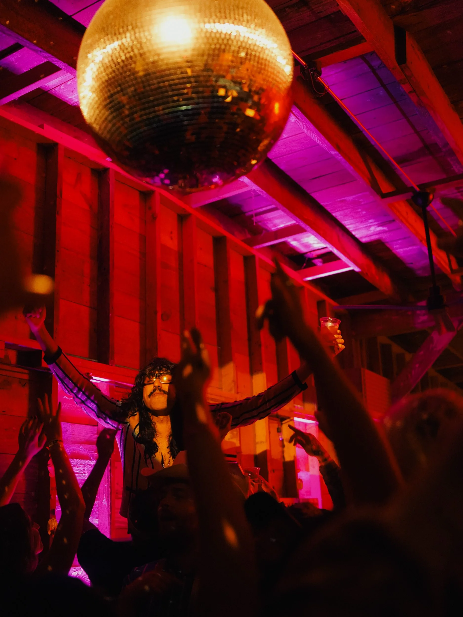 A person with long dark hair, glasses, and a mustache, standing with arms outstretched at a party, surrounded by people with hands raised. A large gold disco ball is hanging from the ceiling, reflecting light. The room has wooden walls and purple lig