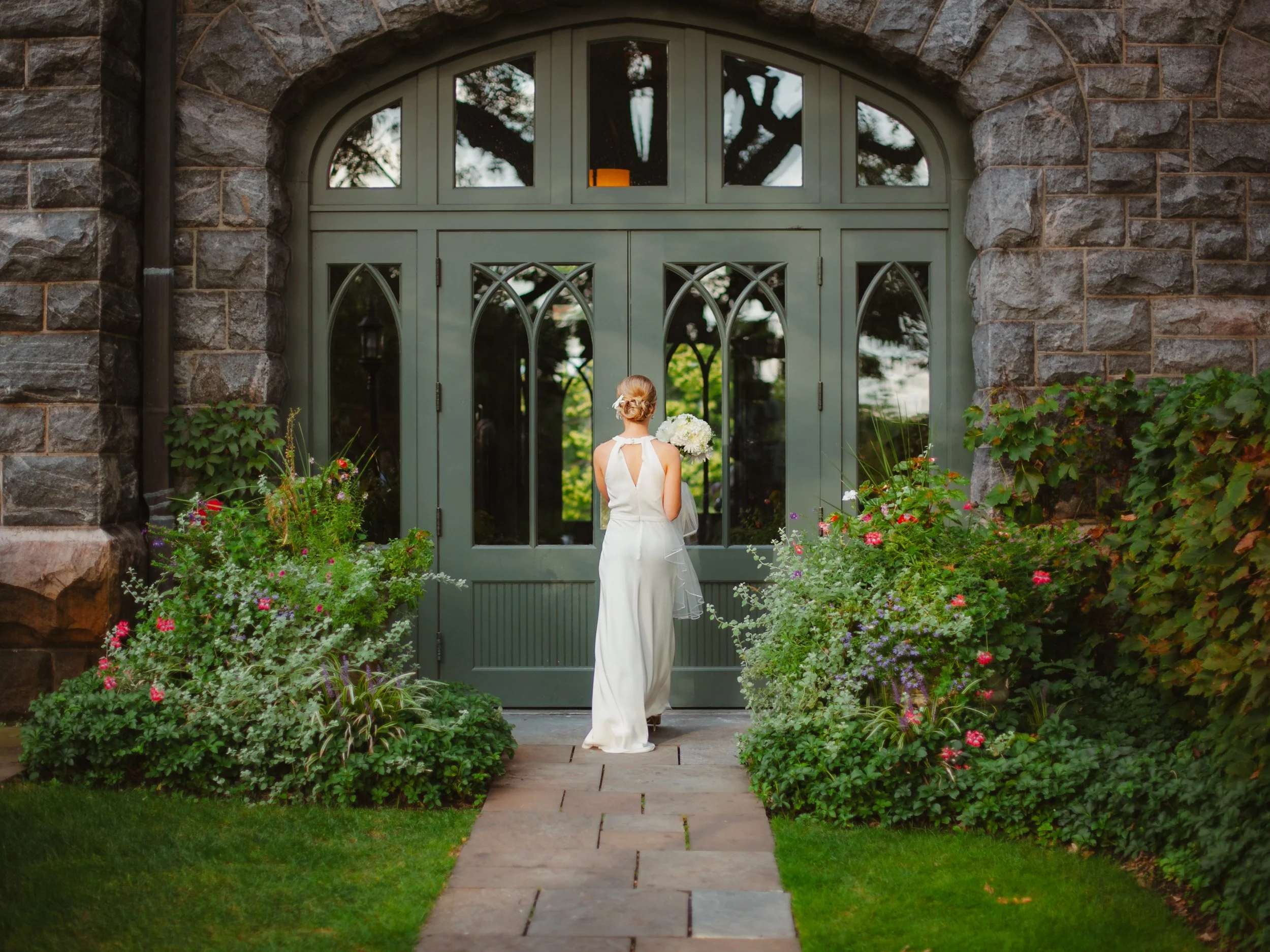 A bride in a white wedding dress with a bouquet standing in front of a large green arched door, surrounded by flower beds and greenery.