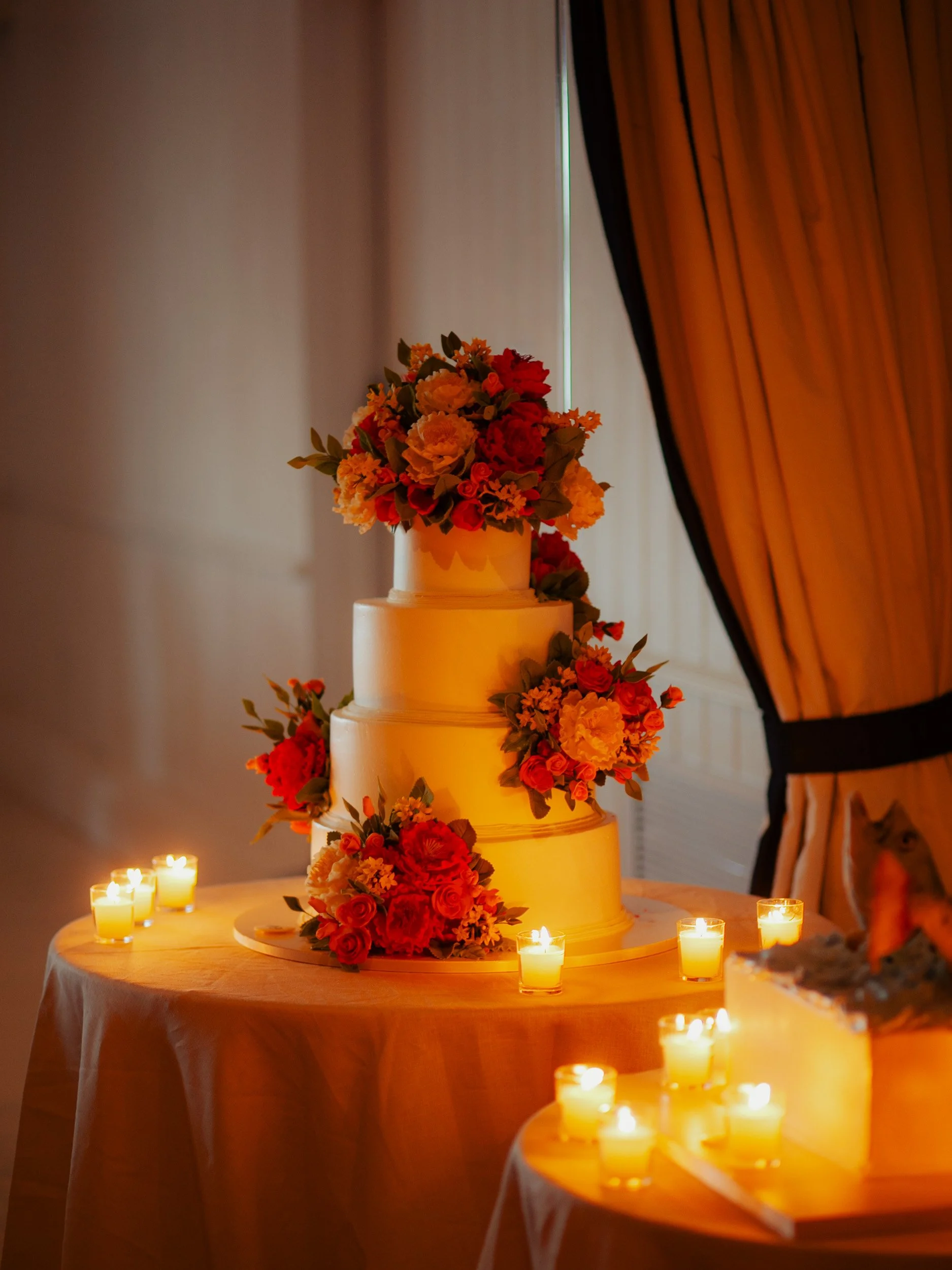 Three-tiered wedding cake decorated with red, orange, and yellow flowers, surrounded by candles, on a table with a white tablecloth, behind a curtain.