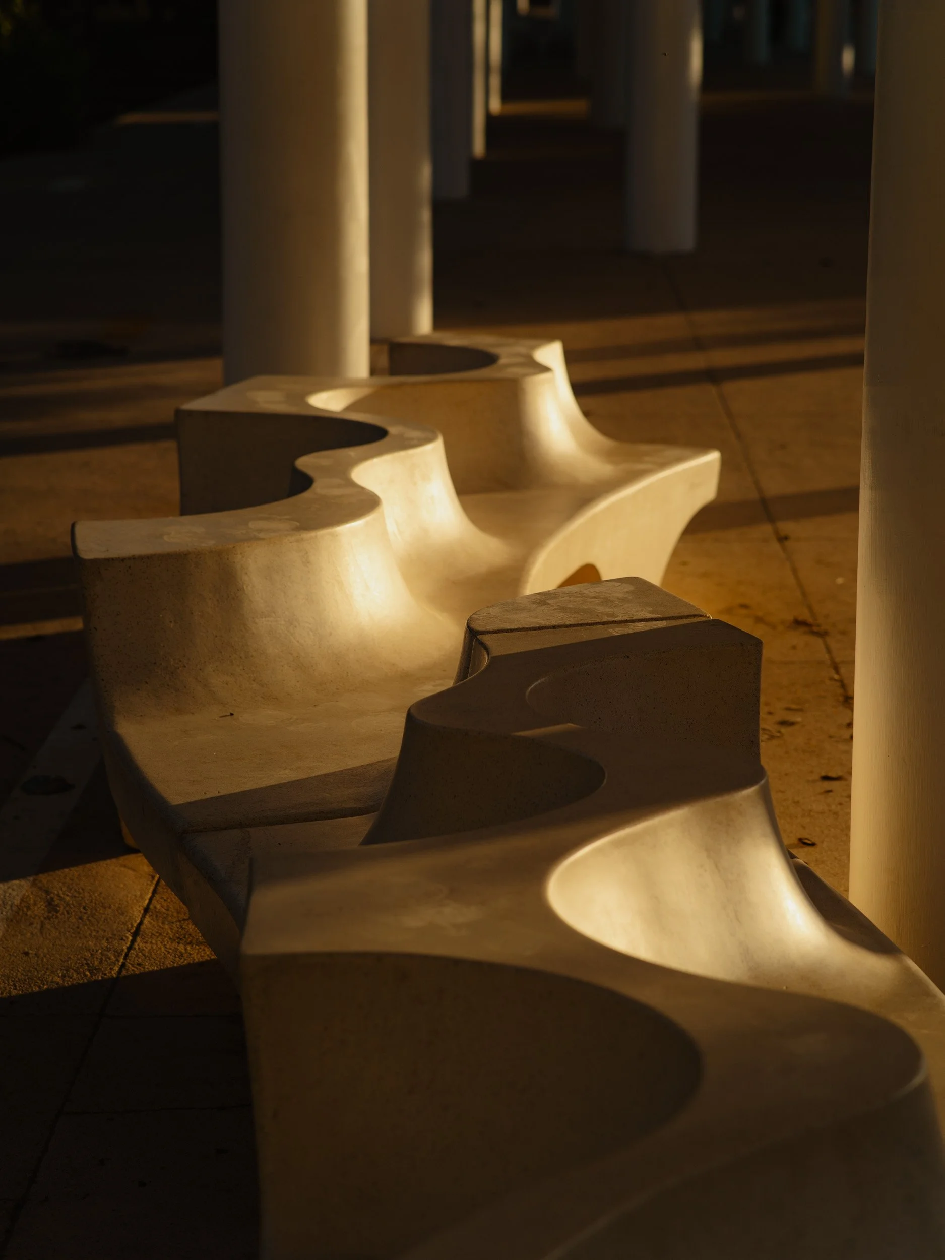 Empty concrete benches illuminated by warm sunlight, arranged in a curve under a pavilion with white columns.