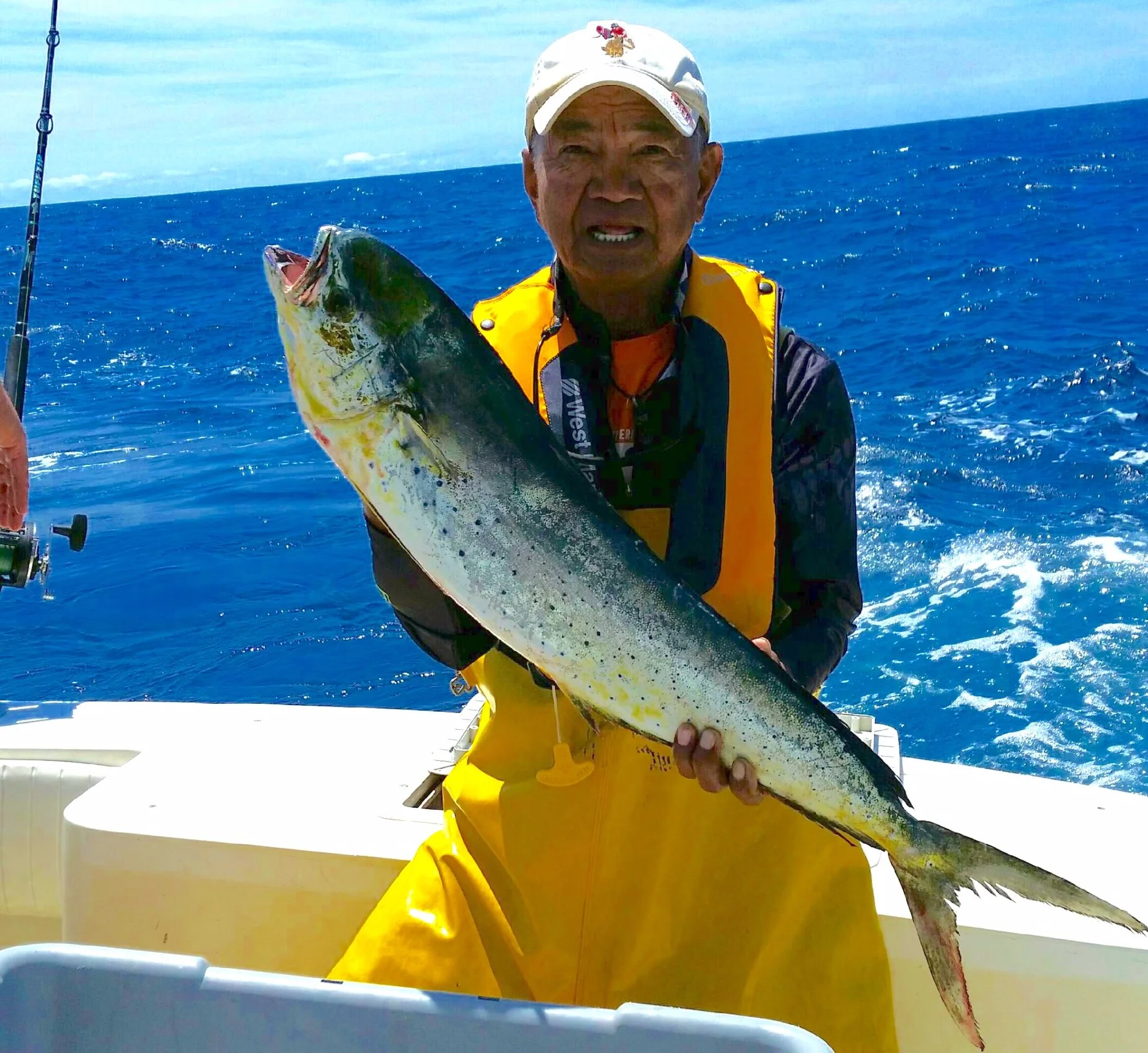 An elderly man in a white cap and yellow waterproof overalls holding a large fish on a boat in the ocean.