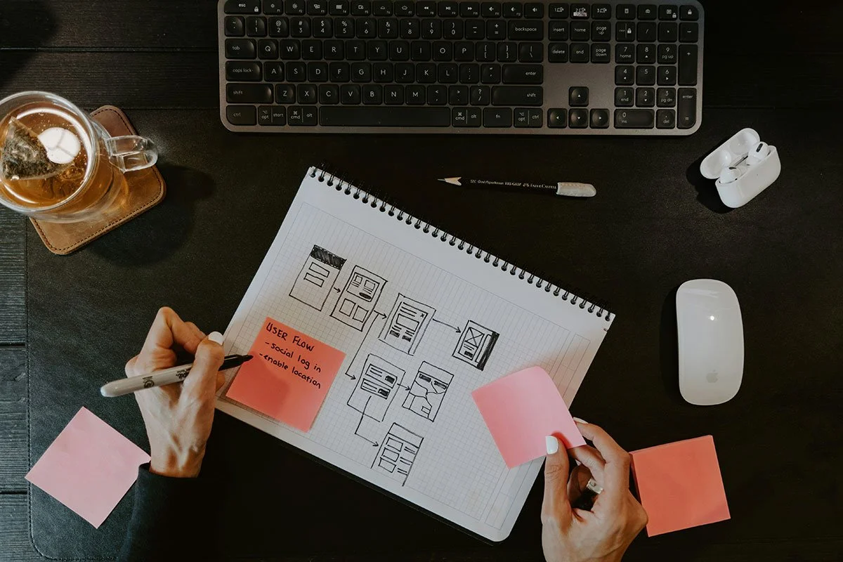 Desk workspace with a computer keyboard, wireless earbuds, a computer mouse, a notepad with sketches and a sticky note, a pen, a person holding a sticky note and writing on it, a glass of tea or beer on a coaster.