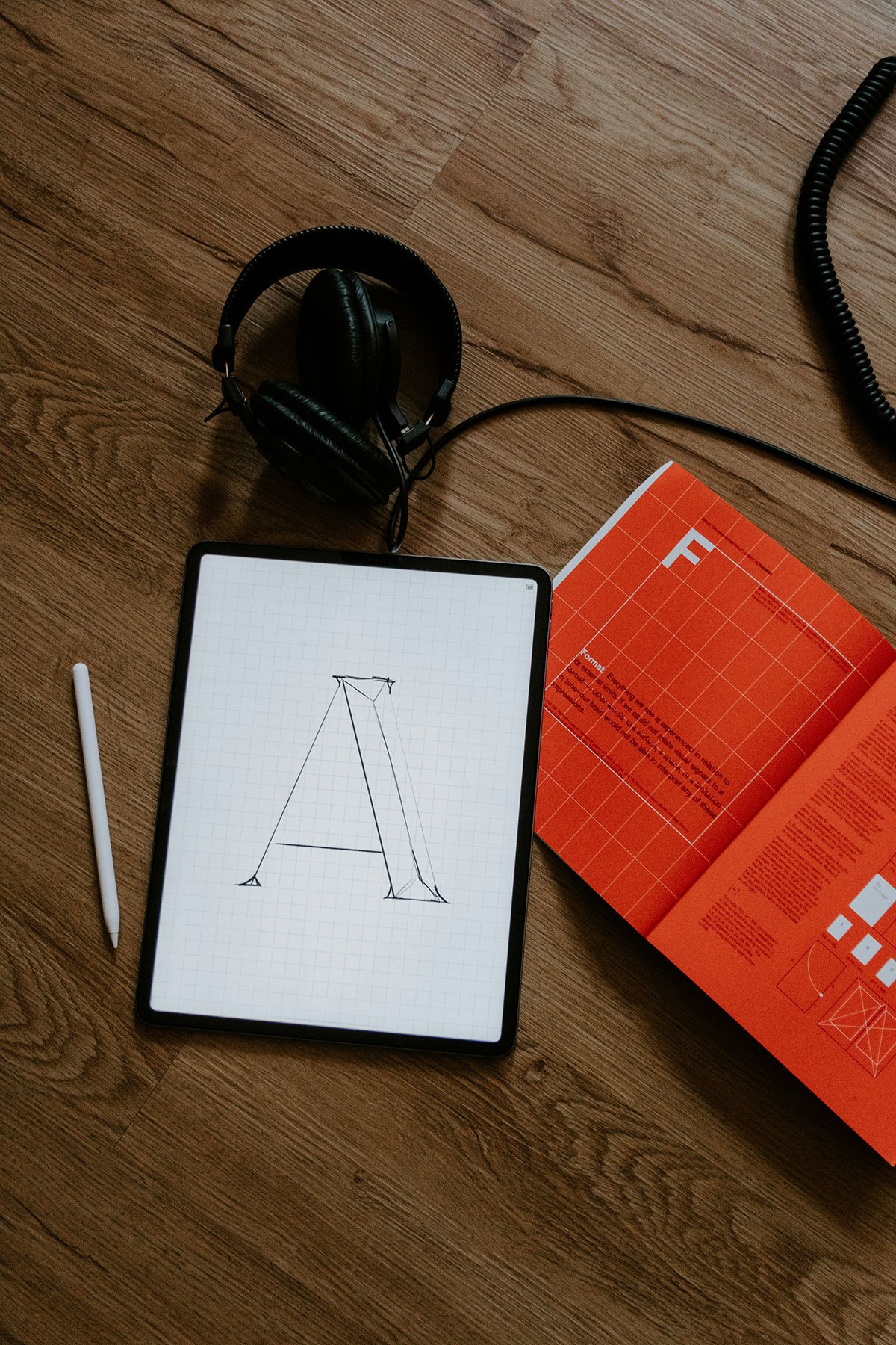 A tablet displaying a sketch of the letter 'A' made of lines. To the left is a white stylus. Above the tablet are black over-ear headphones. To the right is an open orange booklet with text and diagrams on the pages. The setup is on a wooden surface.