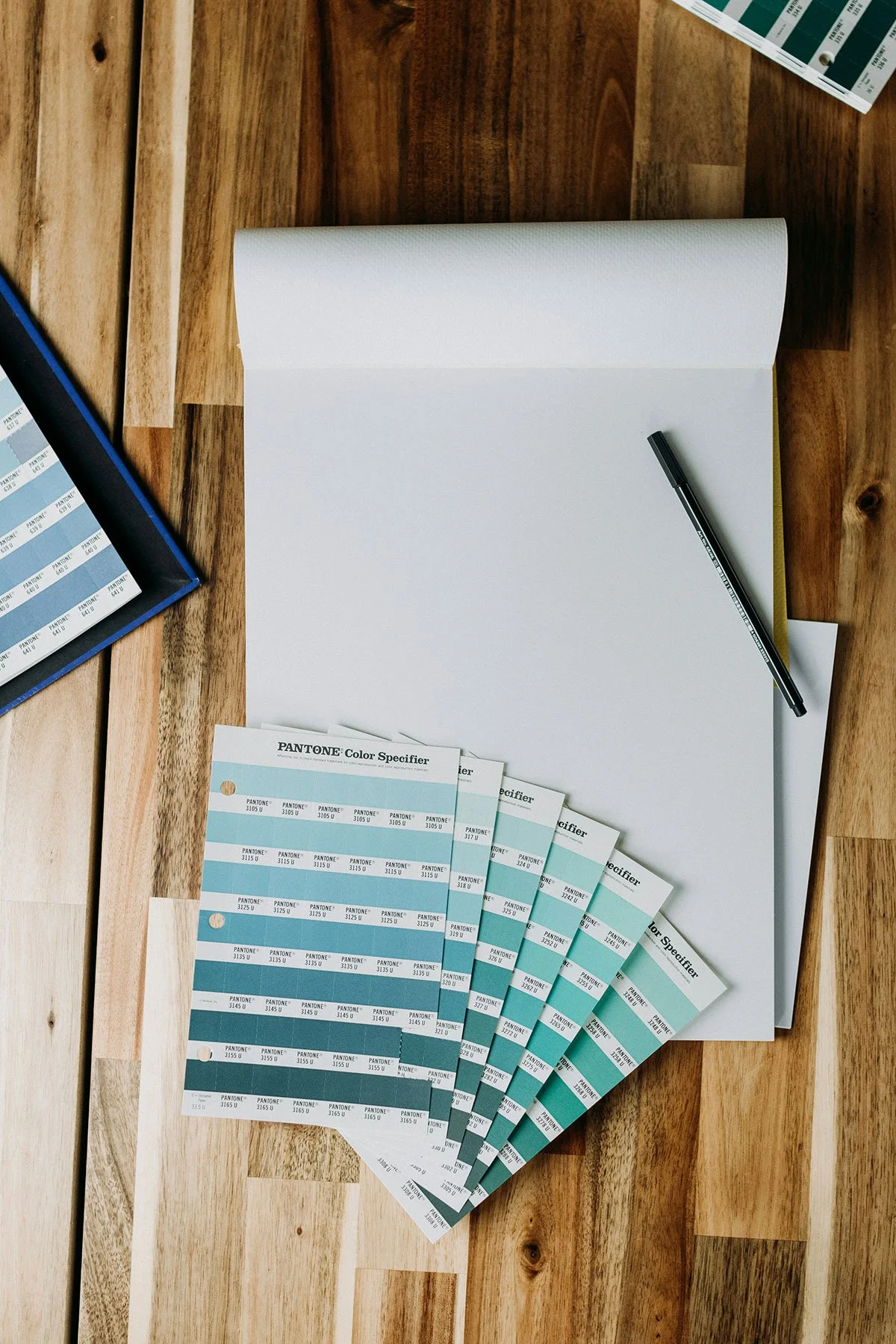 Pantone color swatches in shades of blue and teal spread out on a wooden table next to a white sketchbook, black pen, and color sample sheets.