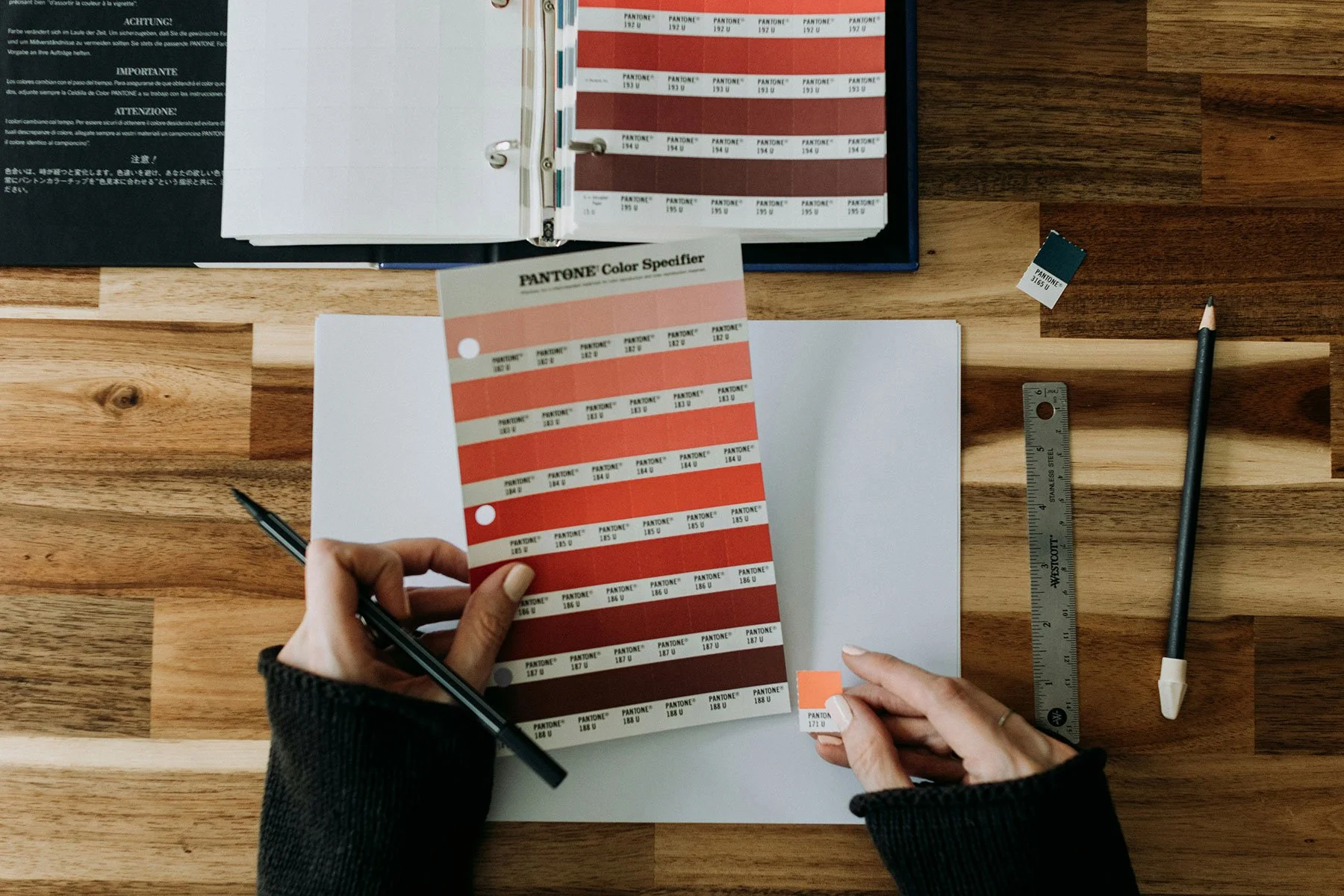 A person holds a Pantone color swatch book, flipping through various shades of orange and red. The person is sitting at a wooden table with other color identification tools, including a ruler, a pencil, and a smaller color swatch. There is an open binder or catalog with additional color swatches in the background.
