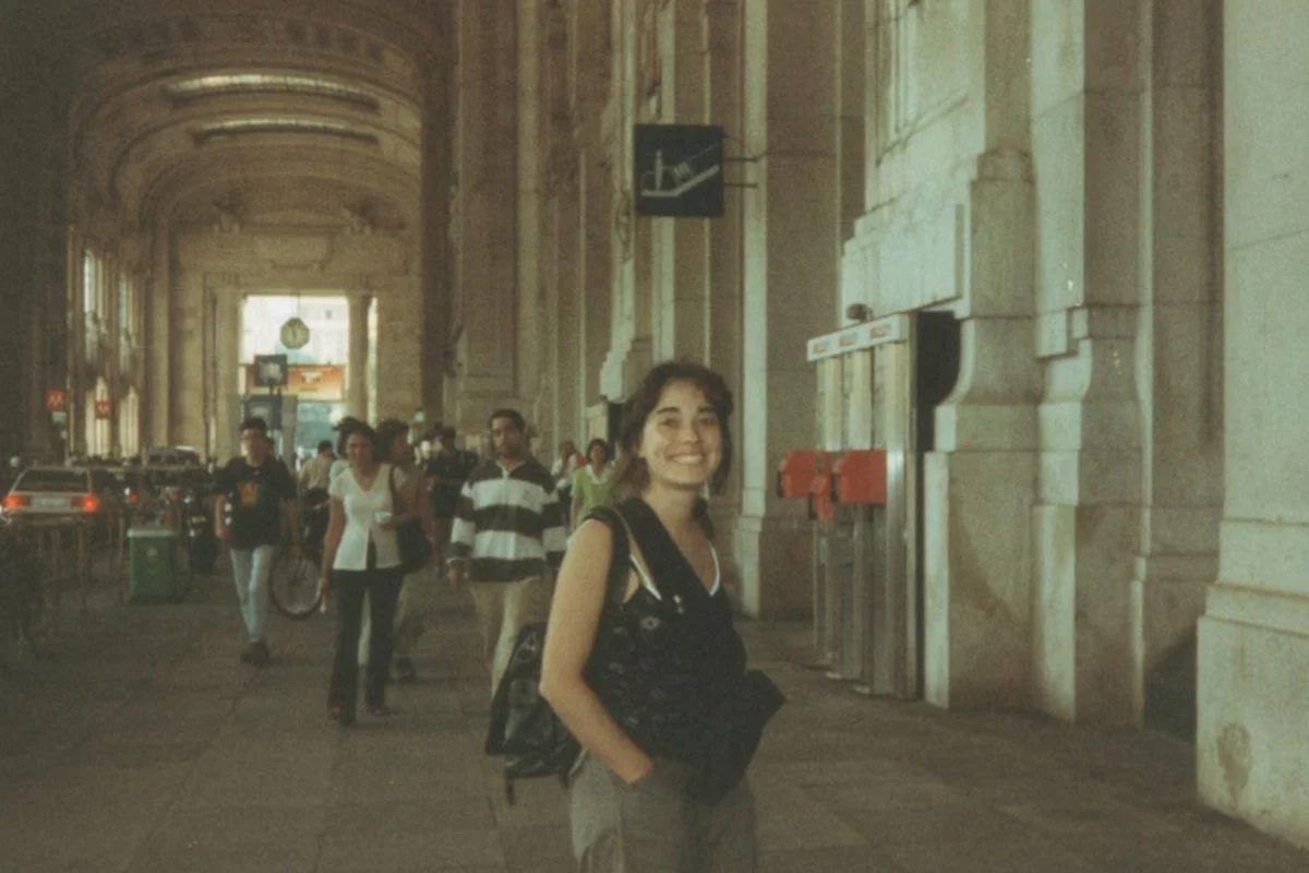 A smiling woman with a backpack and jacket stands inside a large, ornate train station with high arches and other travelers walking in the background.