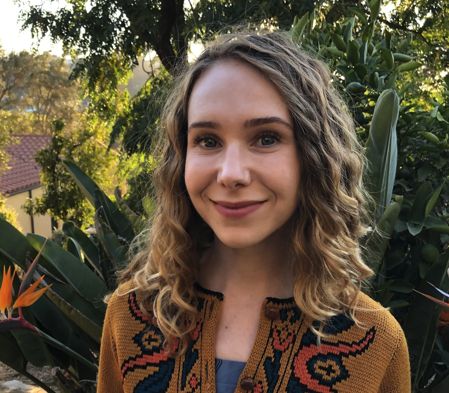 A woman with curly blonde hair and blue eyes smiles outdoors in front of trees and plants, wearing a mustard-colored cardigan with colorful embroidery.