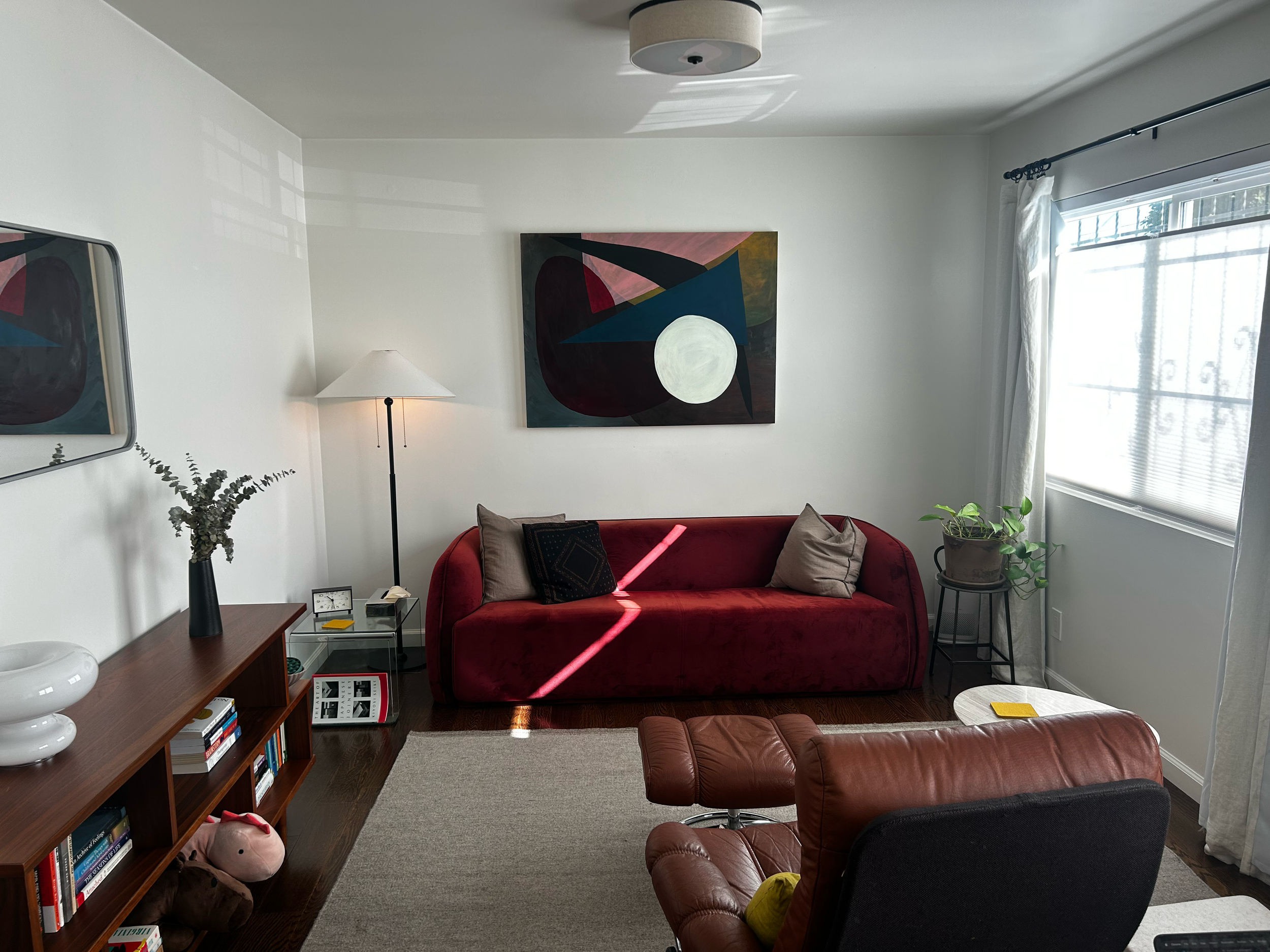 Living room with sunlight coming through window, red velvet sofa with beige and black pillows, wall-mounted abstract art, floor lamp, bookshelf with books and plush toys, potted plant on side table, leather armchair, small round side table, and ceiling light.