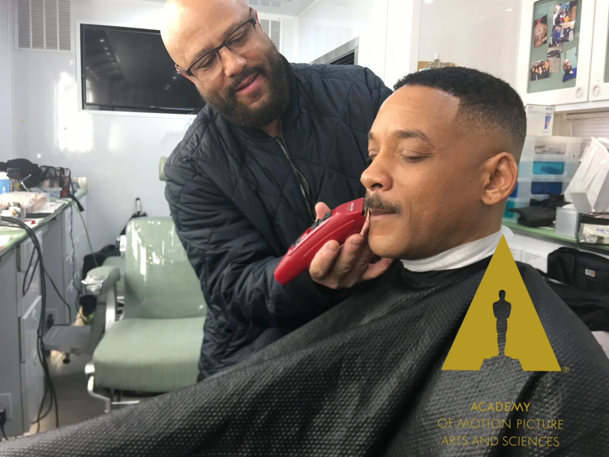 A man getting a close shave with an electric razor by a barber in a salon or clinic. The barber is wearing glasses and a black jacket, and the client has his eyes closed, sitting in a barber chair wrapped in a black cape. The background shows a television, shelves with supplies, and photos on the wall. The photo has a logo for the Academy of Motion Picture Arts and Sciences.