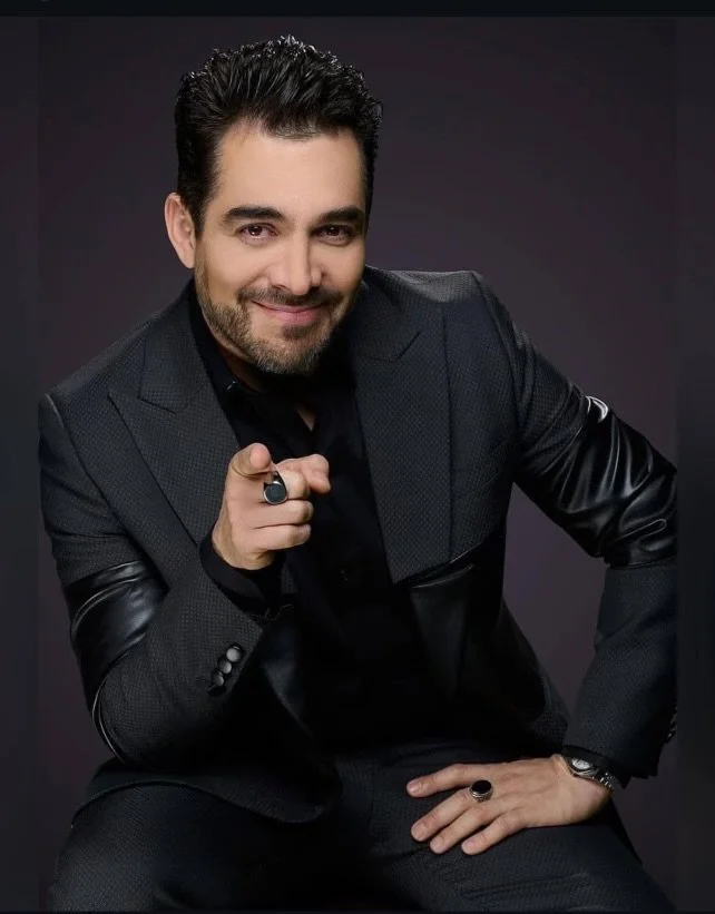 Man with dark hair and beard wearing a black shirt and a dark blazer, pointing at the camera with a smile, seated against a dark background.