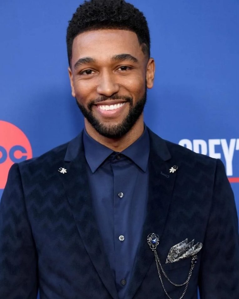 A smiling man with short curly hair and a beard wearing a dark blue suit with a pin on the lapel, a matching shirt, and a decorative pocket square, standing in front of a blue background with some text and a logo.