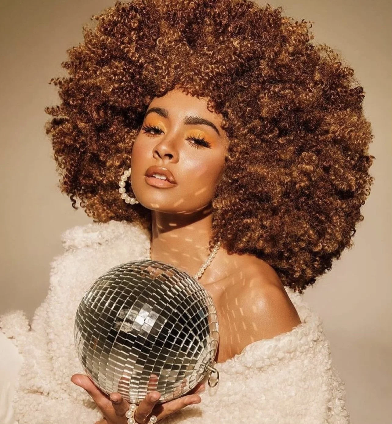 A woman with voluminous curly hair, wearing makeup with orange eyeshadow, holding a disco ball, wearing pearl earrings and a pearl necklace, dressed in a fluffy white garment, and standing against a neutral background.