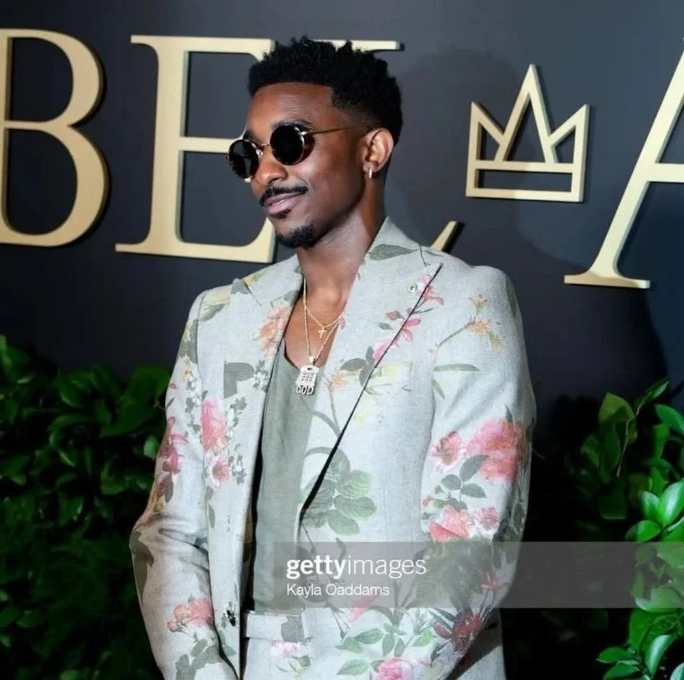 A man wearing sunglasses and a floral suit jacket stands in front of a black background with large letters and a crown logo. There are green plants in the foreground.