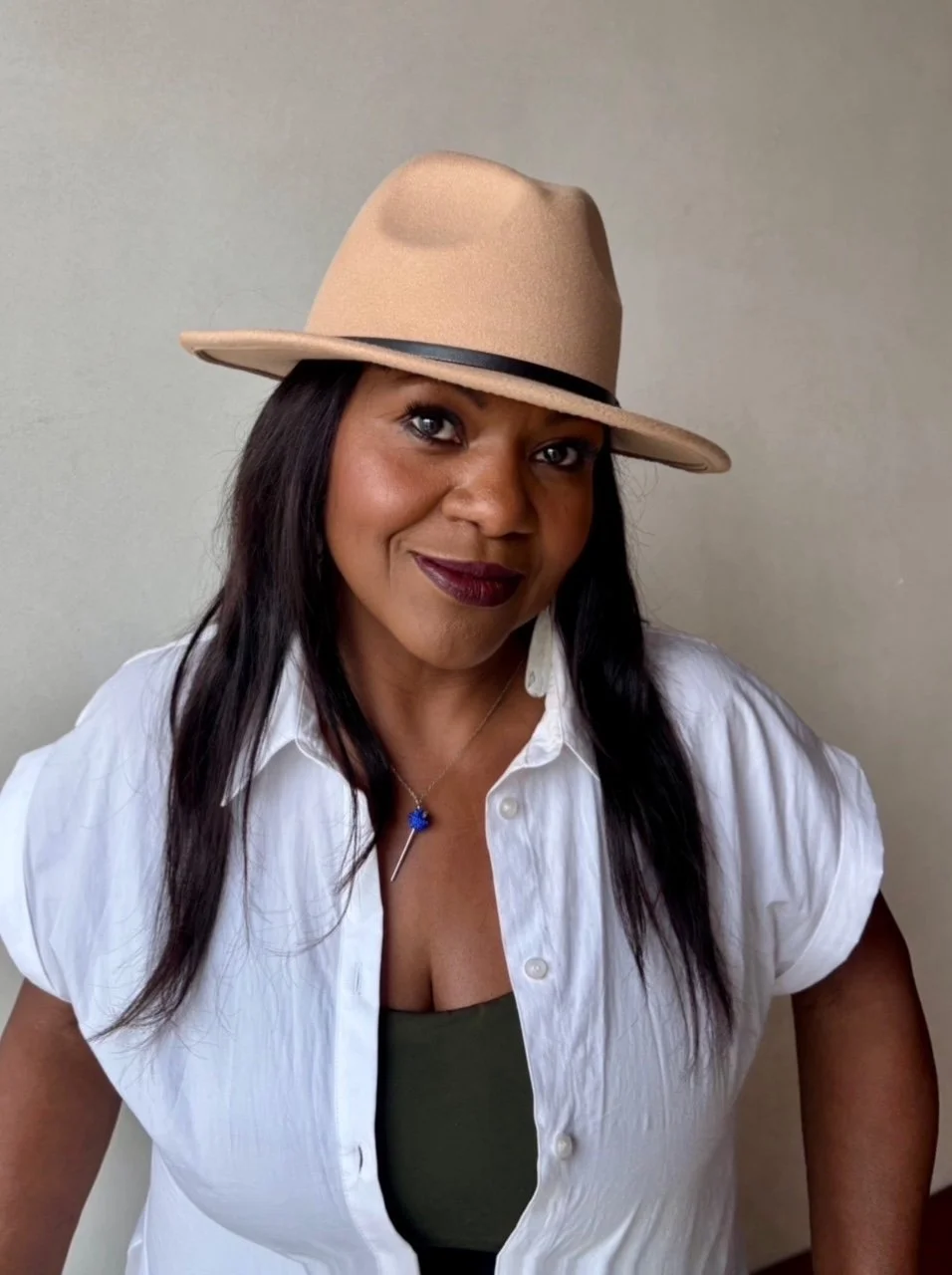 A woman with dark skin and long black hair wearing a beige fedora hat with a black band, a white button-up shirt, a green top underneath, and a necklace with blue and silver pendant, standing against a plain light-colored wall.