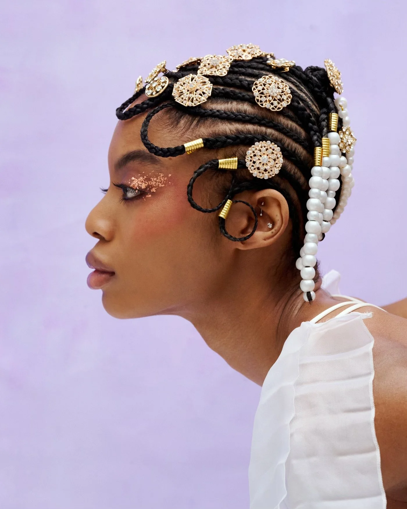 Side profile of a woman with intricately styled braided hair adorned with gold and pearl accessories, wearing makeup with glitter accents on her eyelids, and a white sleeveless top against a soft purple background.