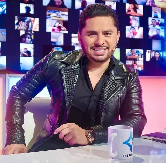 A man with dark hair and a beard, wearing a black leather jacket, sitting at a table with a white mug, in a TV studio with a background of multiple screens displaying video feeds.