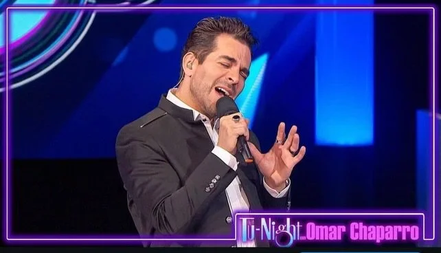 A man wearing a dark suit and white shirt singing into a microphone on stage with neon lights and a sign reading 'L Night Omar Chaparro' in the background.