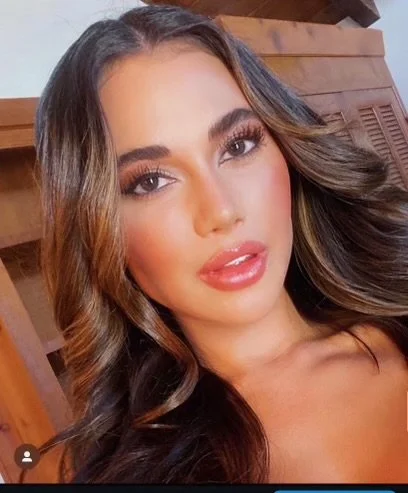 Close-up of a woman with long, wavy brown hair, wearing makeup, and looking at the camera. Background features wooden furniture.