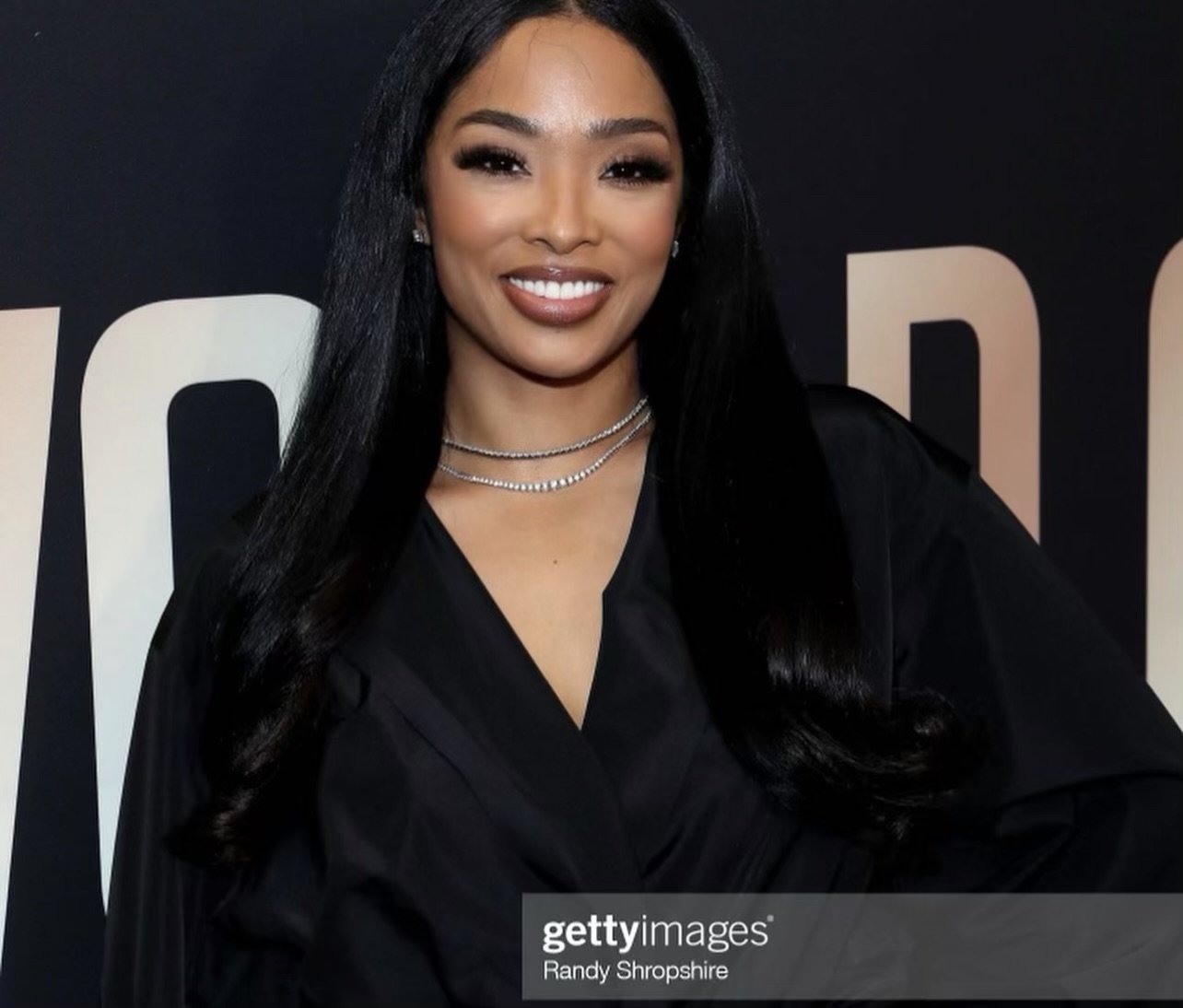 Woman with long black hair smiling, wearing layered necklaces and a black outfit, standing in front of a dark backdrop with partial text.