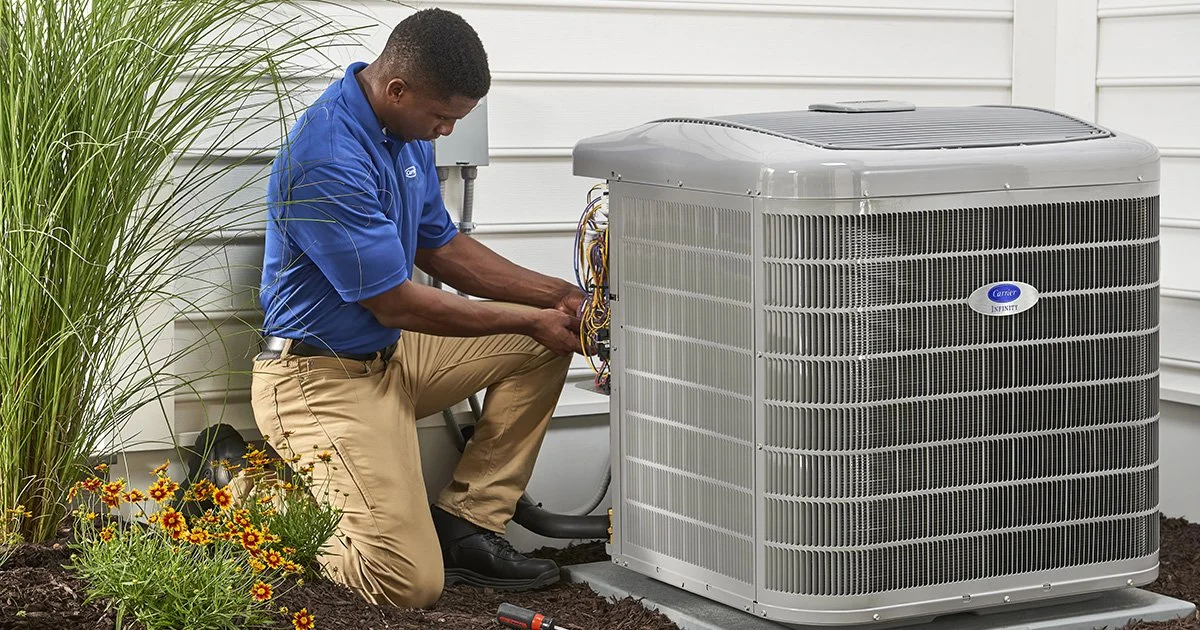 Technician working on a large outdoor air conditioning unit outside a house with white siding, next to a garden with flowers and tall grass.