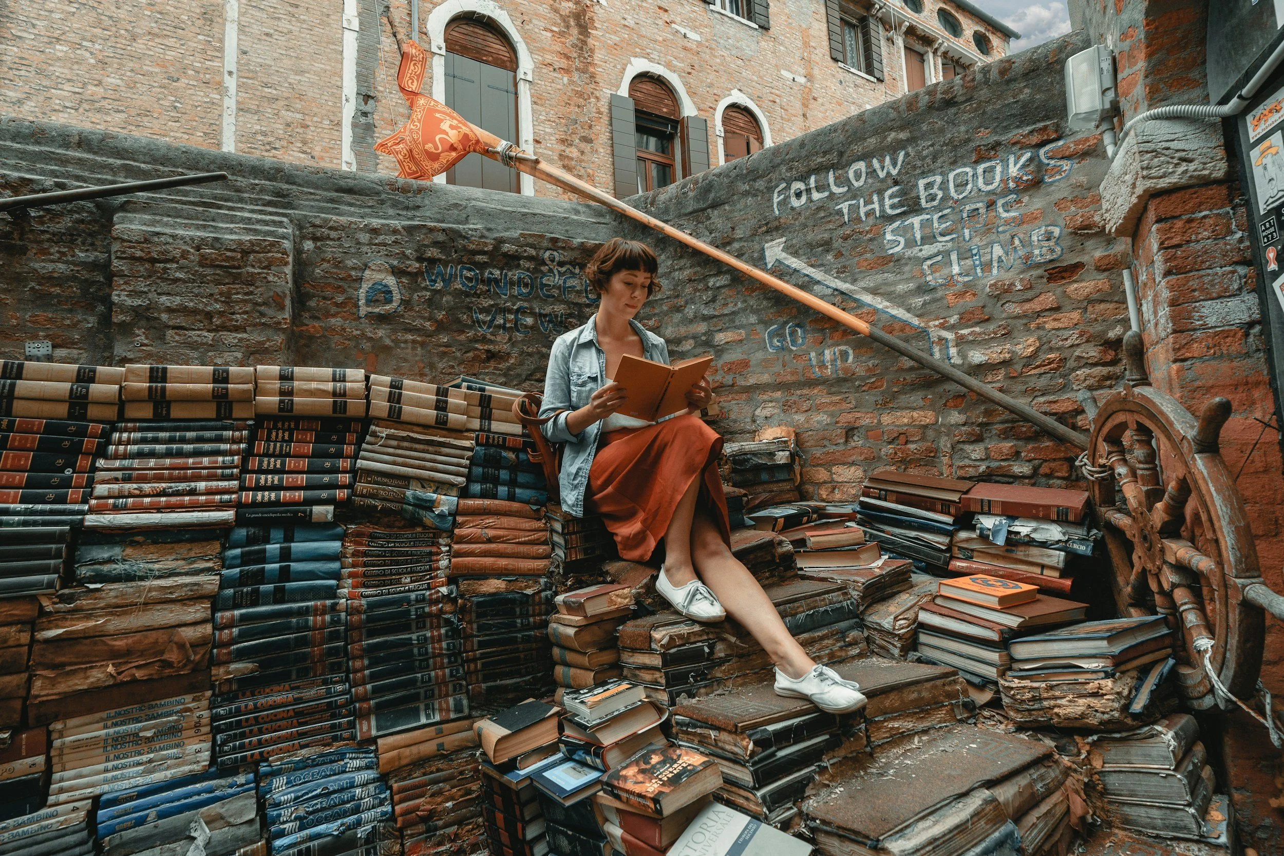 A woman sitting on a pile of old books and reading a book in an outdoor setting with a brick wall background