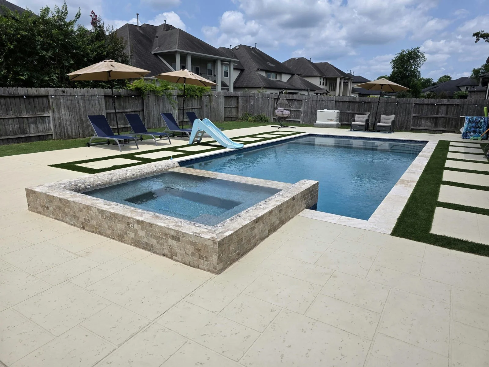 Backyard swimming pool with a hot tub, surrounded by lounge chairs, umbrellas, and outdoor furniture, enclosed by a wooden fence, with houses and a cloudy sky in the background.