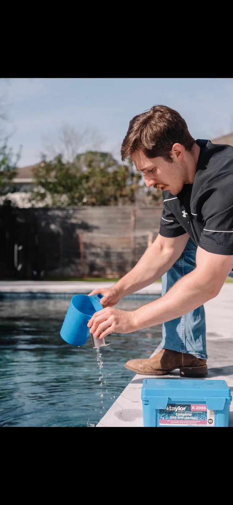 Man pouring pool chemicals into a backyard swimming pool from a blue container.