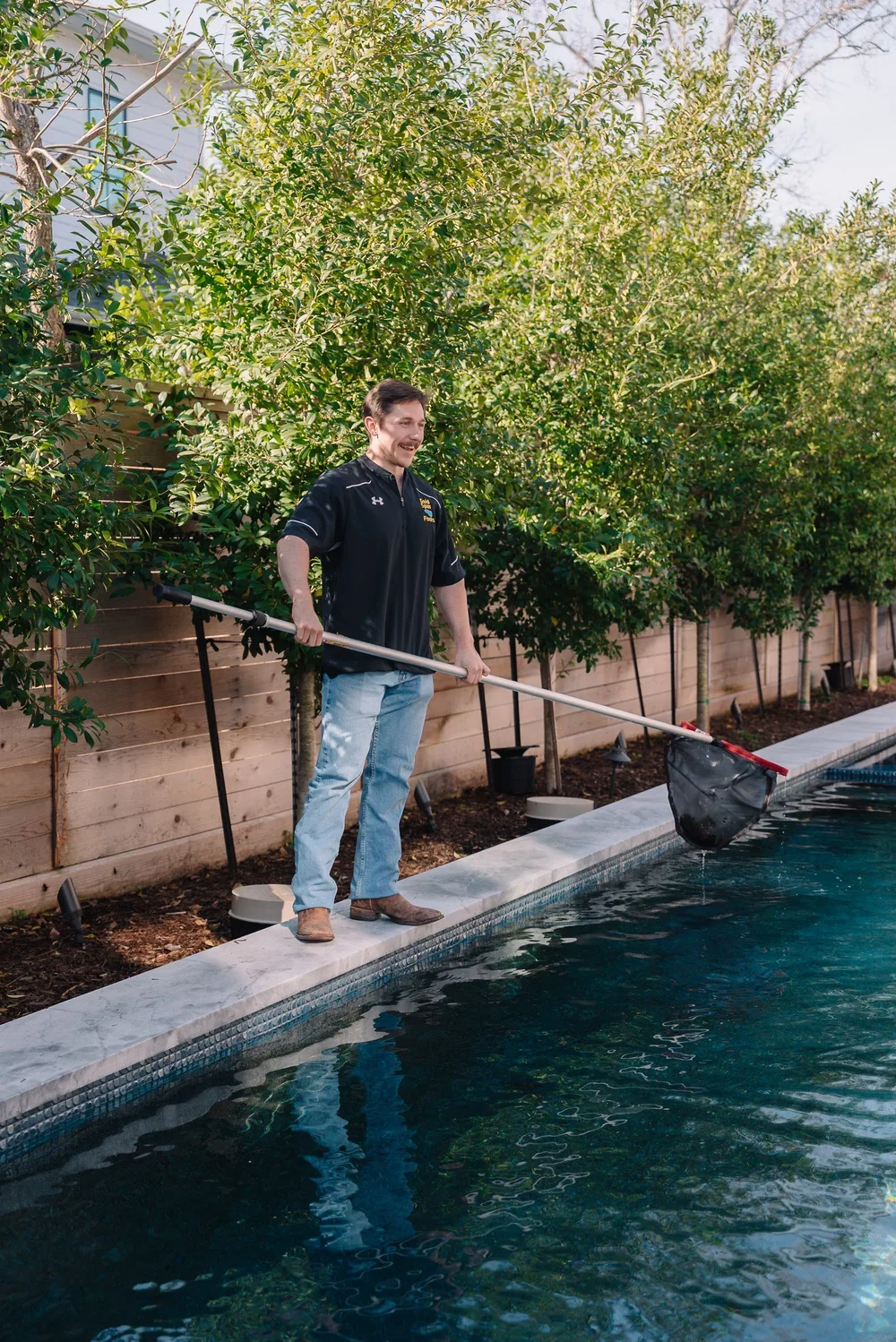 A man standing on the edge of a pool, holding a pool skimmer net, with trees and a wooden fence in the background.