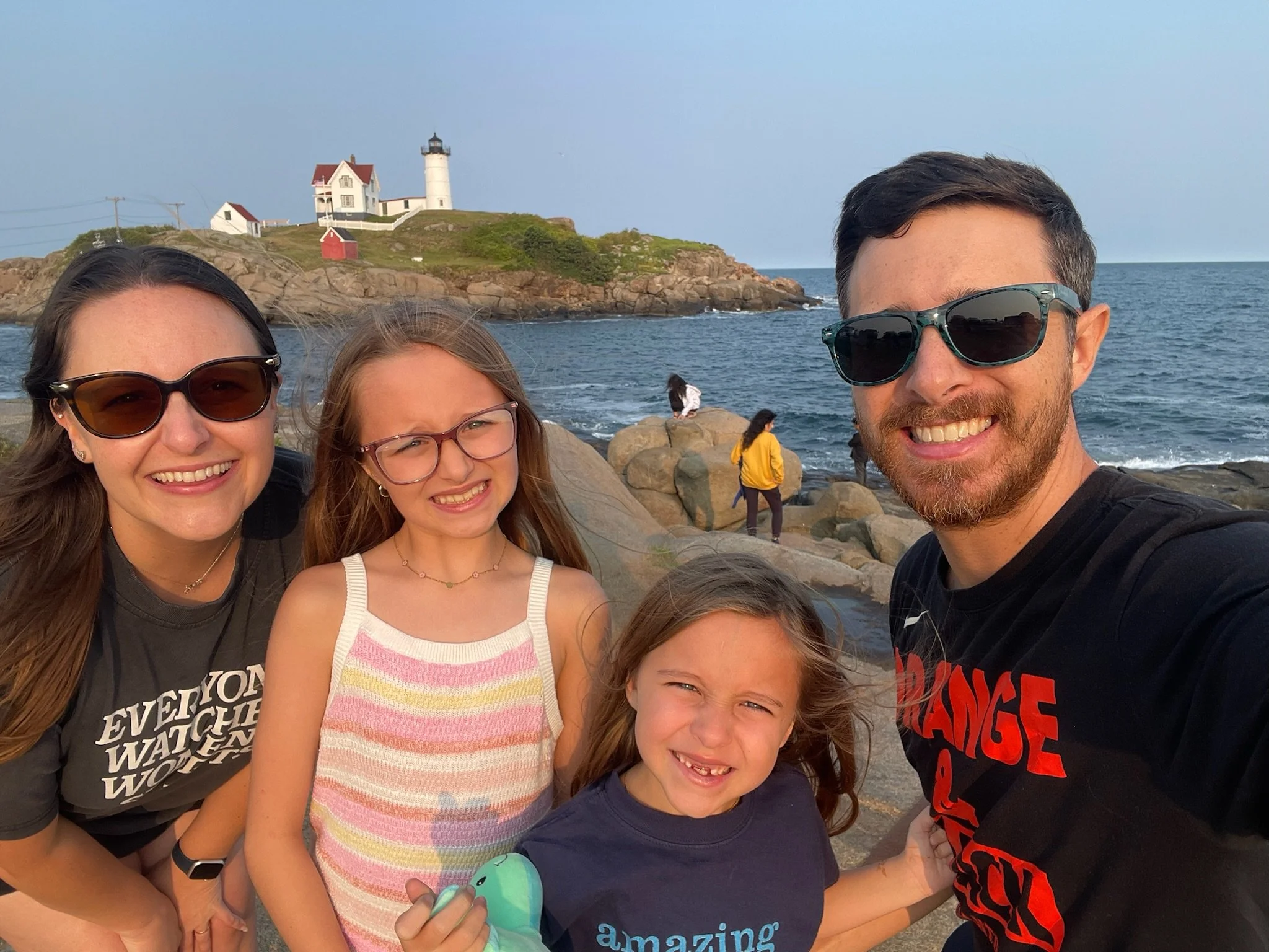 A family of four taking a selfie on rocky terrain by the ocean, with a lighthouse and a house on a hill in the background. The family members are smiling, wearing casual clothes and sunglasses.