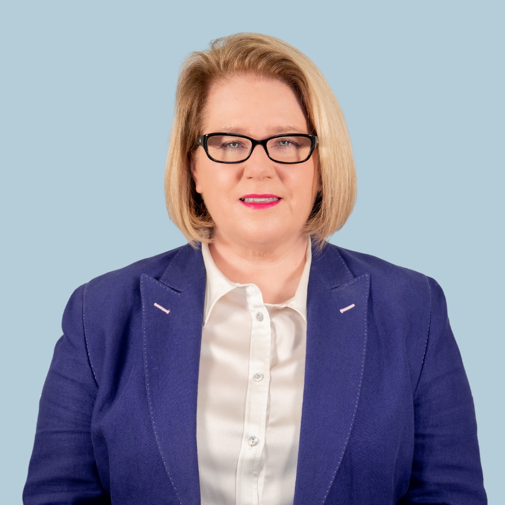 A woman with shoulder-length curly dark hair, wearing a white blazer over a bright yellow top, stands against a solid purple background with her arms crossed and a slight smile on her face.