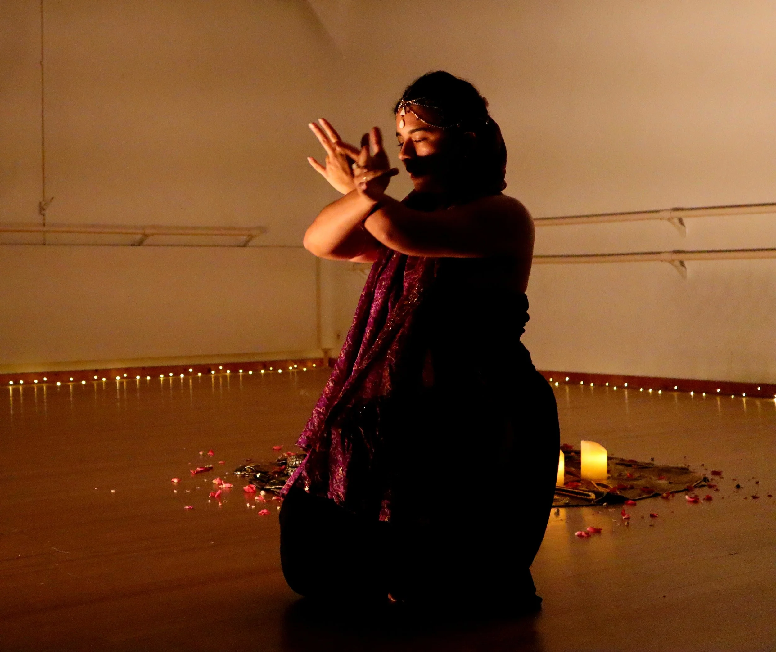 Une femme en tenue bohème danse dans une pièce tamisée, décorée de bougies, de pétales de fleurs et de petites lumières. Elle porte une robe longue et un bandeau pour la tête.