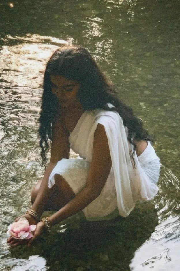 Femme en robe blanche agenouillée dans une rivière, tenant une fleur rose dans ses mains.