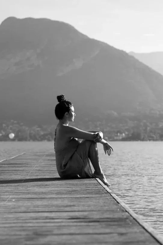 Une femme assise sur un quai en bois, regardant l'eau, avec une montagne en arrière-plan.