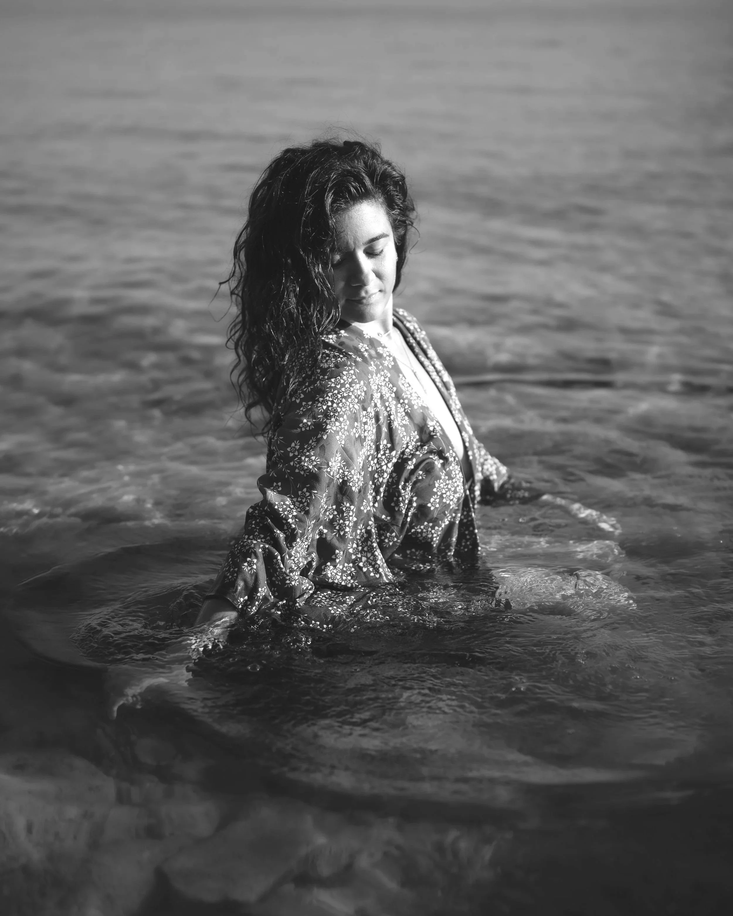 Une femme avec des cheveux bouclés dans l'eau de la mer, portant une robe ou un vêtement avec un motif floral, yeux fermés, visage paisible.