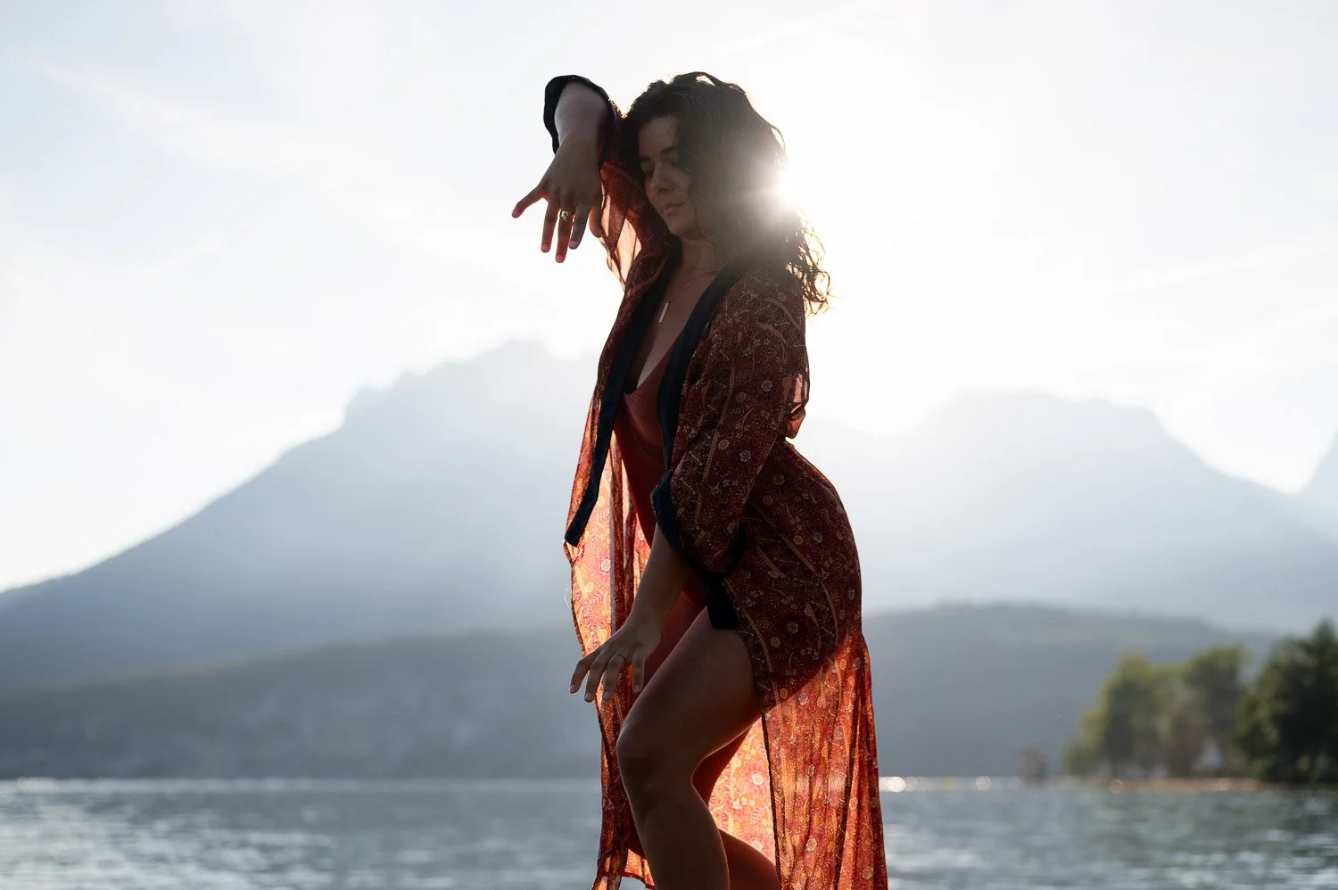 Femme posant devant un paysage de montagne et d'eau au coucher du soleil, portant une robe longue et transparente.