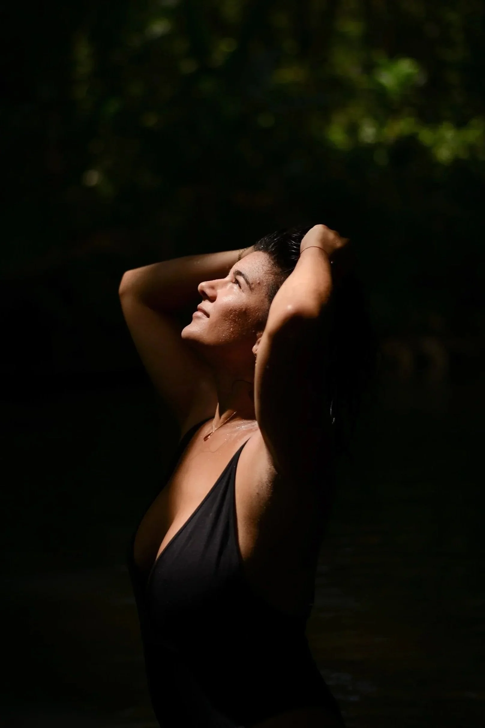 Femme souriante avec les bras levés, portant un maillot de bain noir, se tenant dans un environnement naturel à l'ombre avec un éclairage solaire.