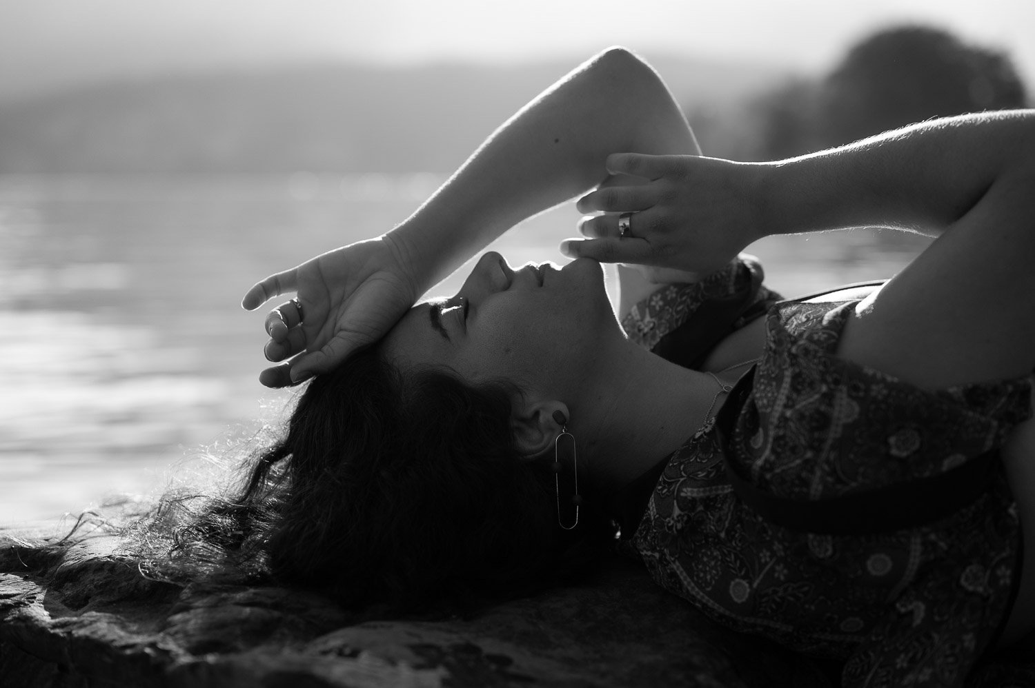 Femme allongée sur le dos près de l'eau, portant un vêtement à motifs, les yeux fermés, avec une main reposant sur son front et une boucle d'oreille à une oreille, en noir et blanc.