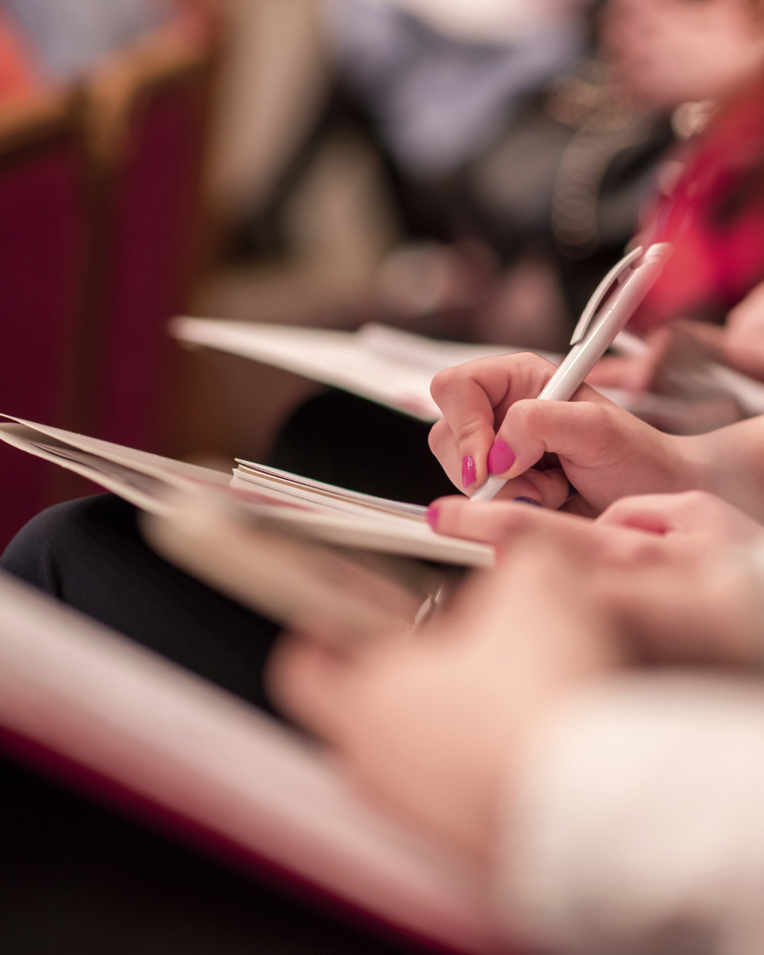 Main tenant écrivant dans un carnet avec un stylo blanc, avec d'autres personnes assises en arrière-plan.