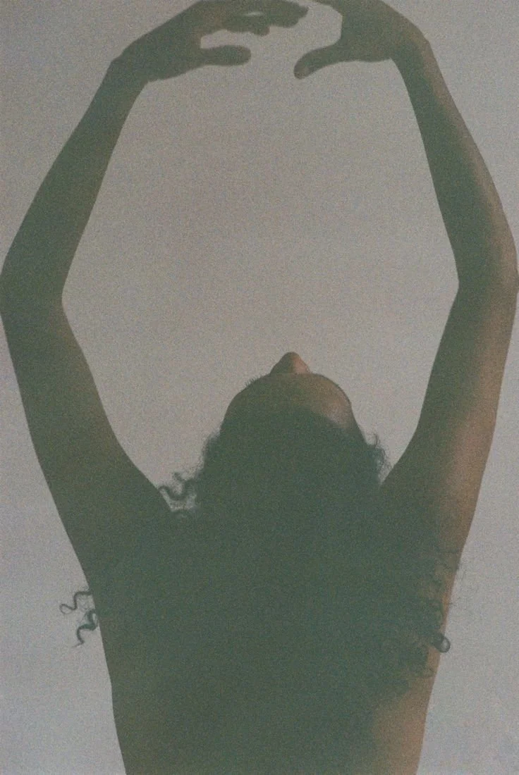 Une silhouette de personne vue de dessous, avec les bras levés au-dessus de la tête, dans un espace obscur ou faiblement éclairé.