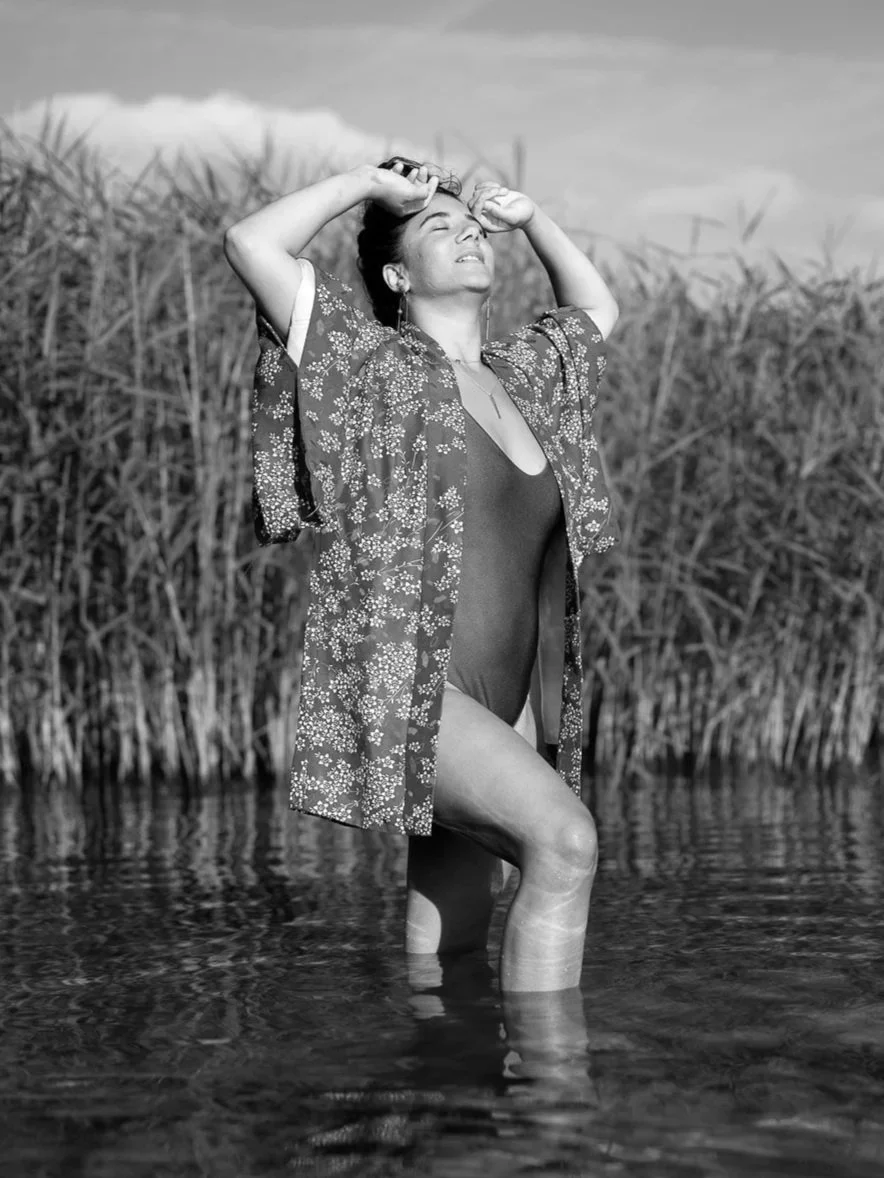 Une femme souriante en maillot de bain et robe légère, debout dans l'eau, avec des herbes hautes en arrière-plan.