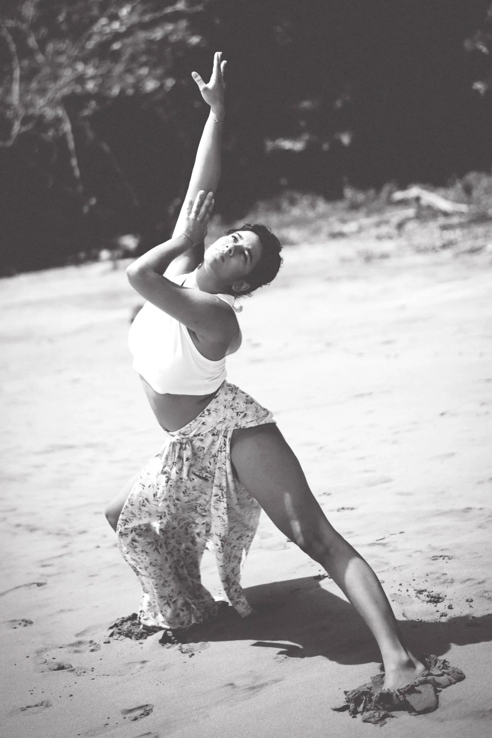 Femme qui pose une danse expressive sur la plage, en costume léger, avec des dunes et des arbres en arrière-plan.