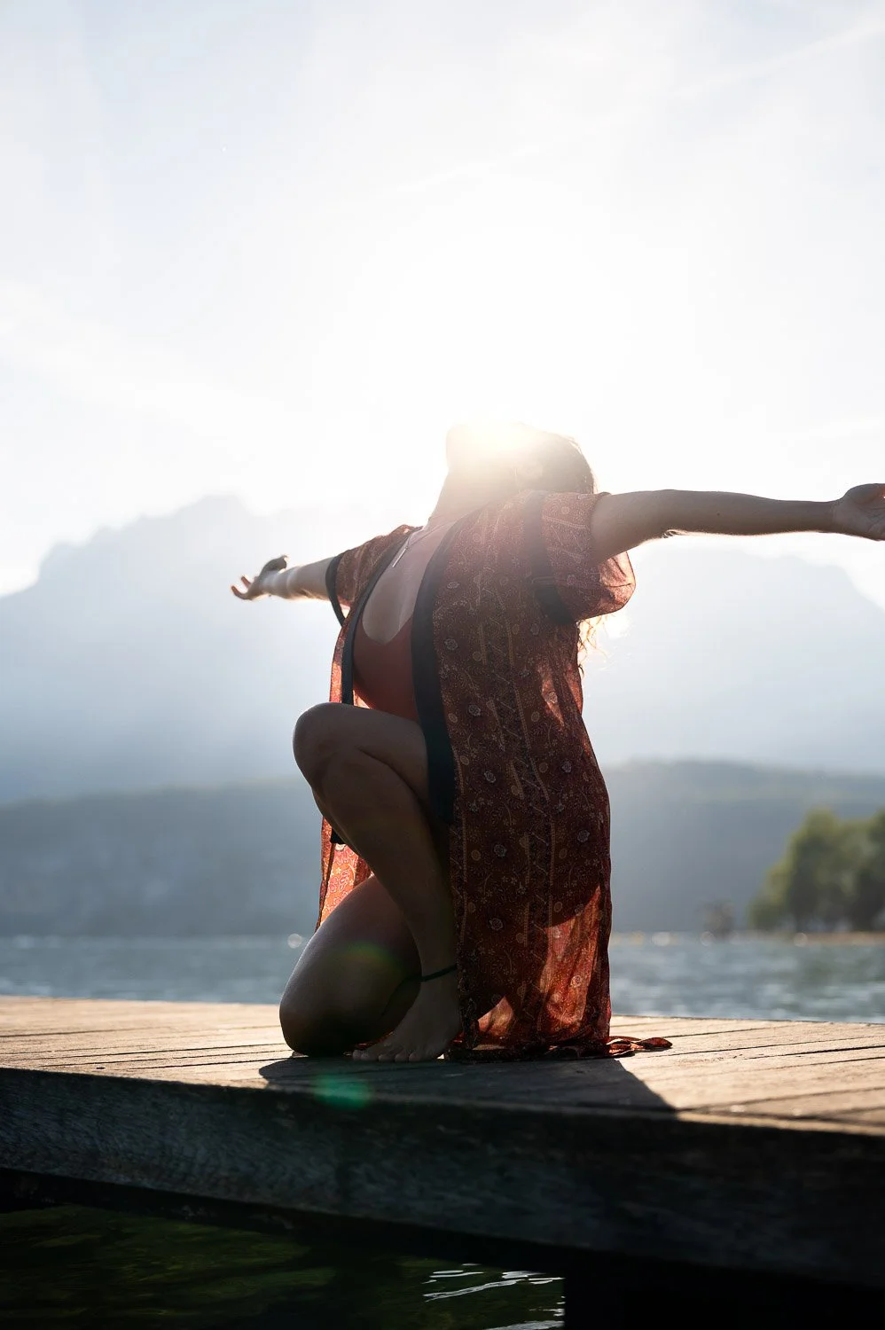 Femme faisant du yoga en posture de guerrier sur un pont en bois au bord d'un lac, avec des montagnes en arrière-plan, au coucher du soleil.