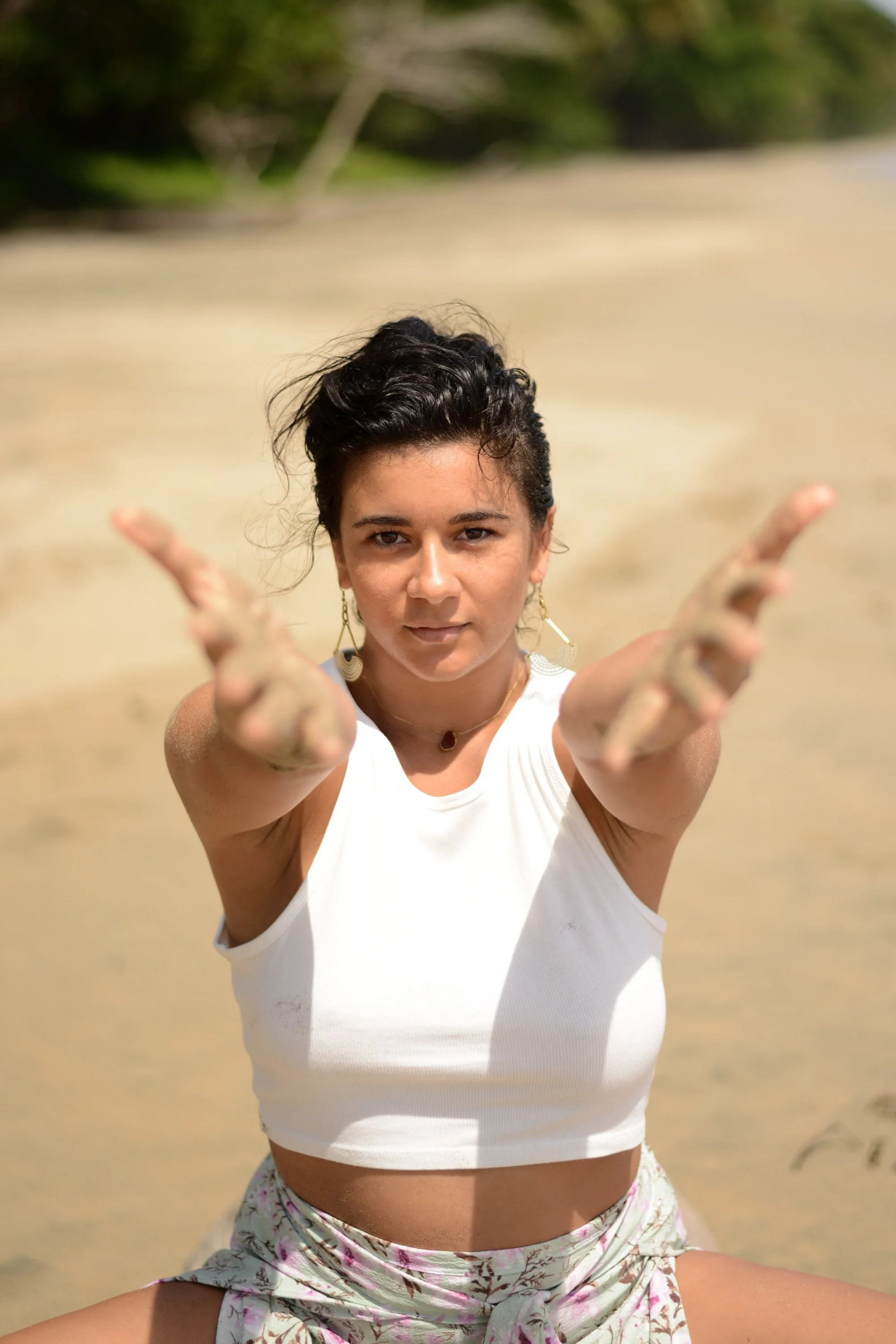 Jeune femme sur la plage, regardant l'appareil photo, avec les bras tendus vers lui, cheveux bouclés courts, portant des boucles d'oreilles pendantes, une chaîne avec un pendentif, et un haut blanc, en train de poser pour une photo.