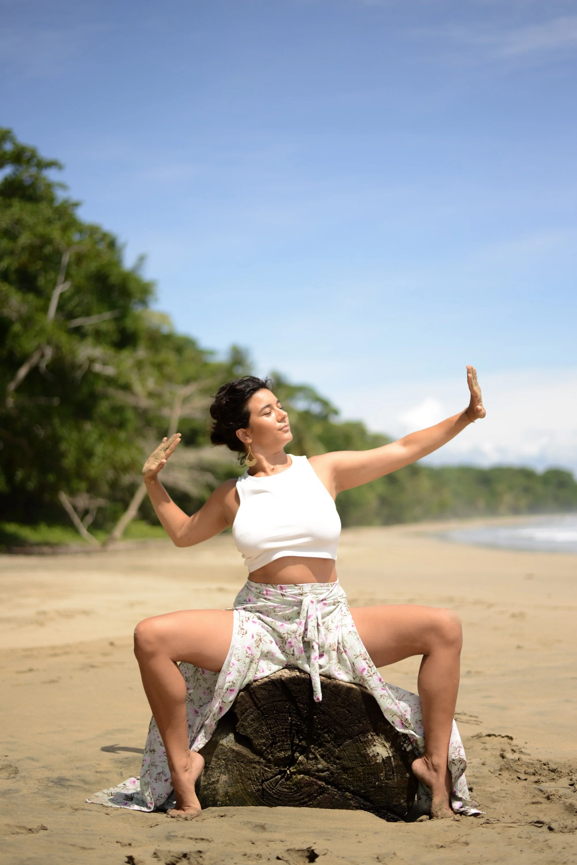 Une femme en posture de méditation ou de yoga sur une plage, assise sur un morceau de bois, avec la mer et la forêt en arrière-plan, le ciel est clair et ensoleillé.