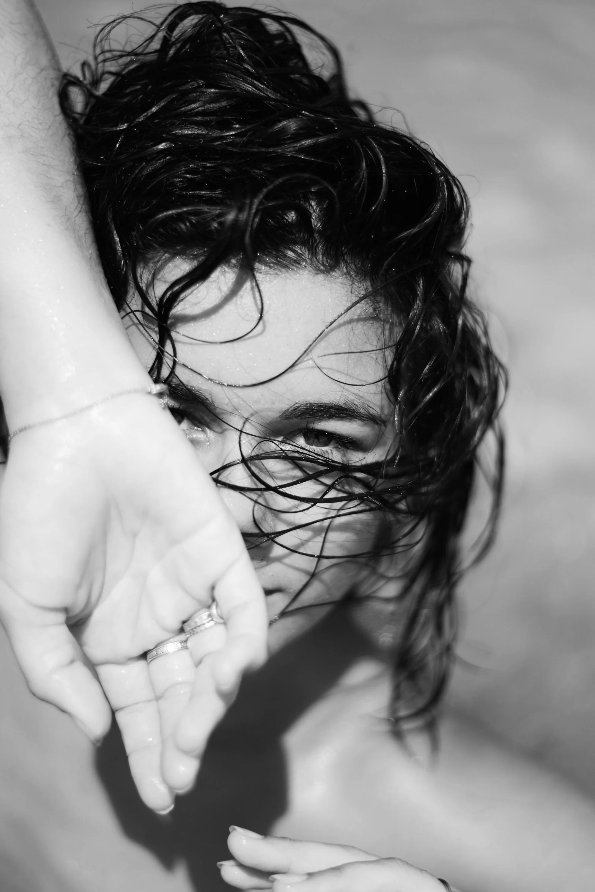 Une femme aux cheveux mouillés, partiellement cachée derrière sa main, regarde intensément la caméra, en noir et blanc.