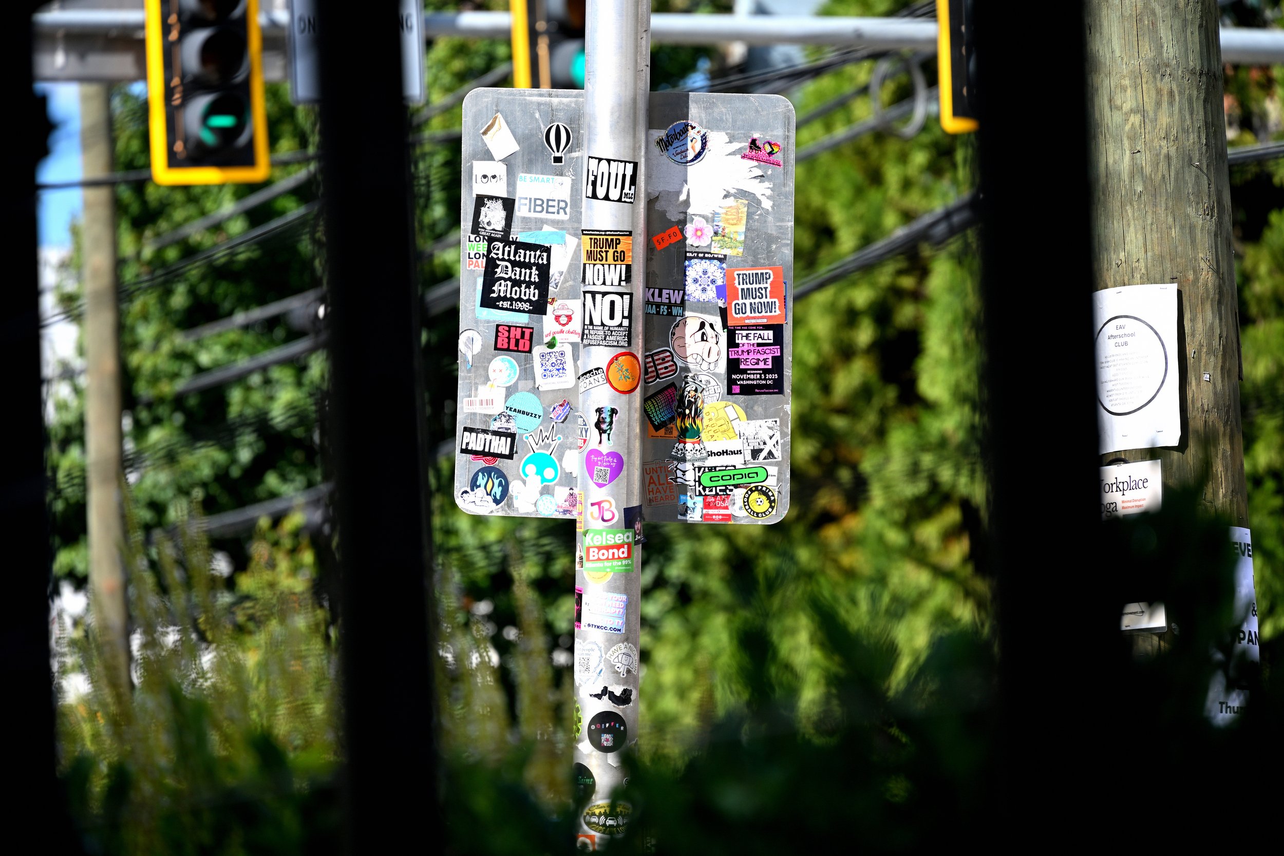 A metal signpost covered with numerous stickers, located outdoors behind some wooden and metal fence posts, with trees and foliage in the background.