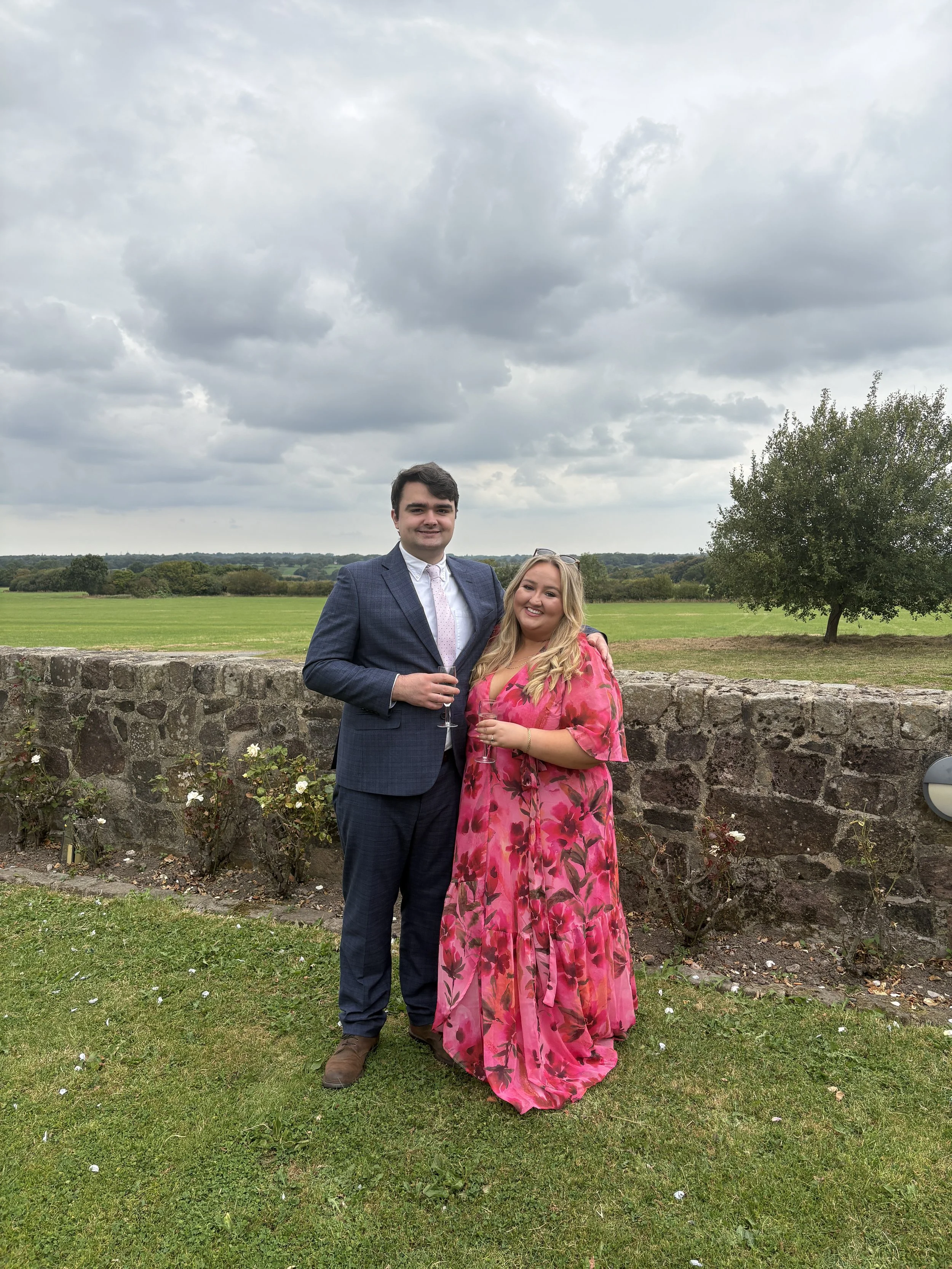A man in a dark blue suit and a woman in a pink floral dress standing outdoors on a grassy area, holding champagne glasses, with a stone wall, green field, and cloudy sky in the background.
