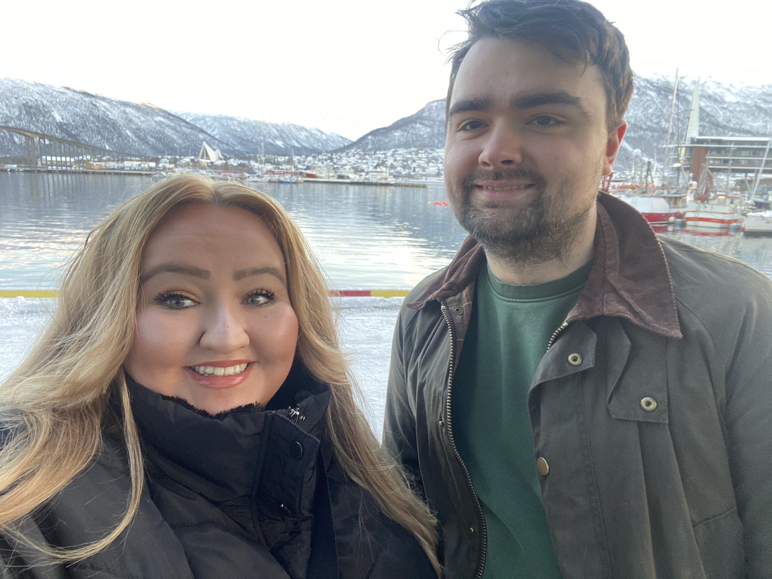 A smiling woman with long blonde hair and a man with dark hair and a beard standing near a harbor with snow-capped mountains in the background.